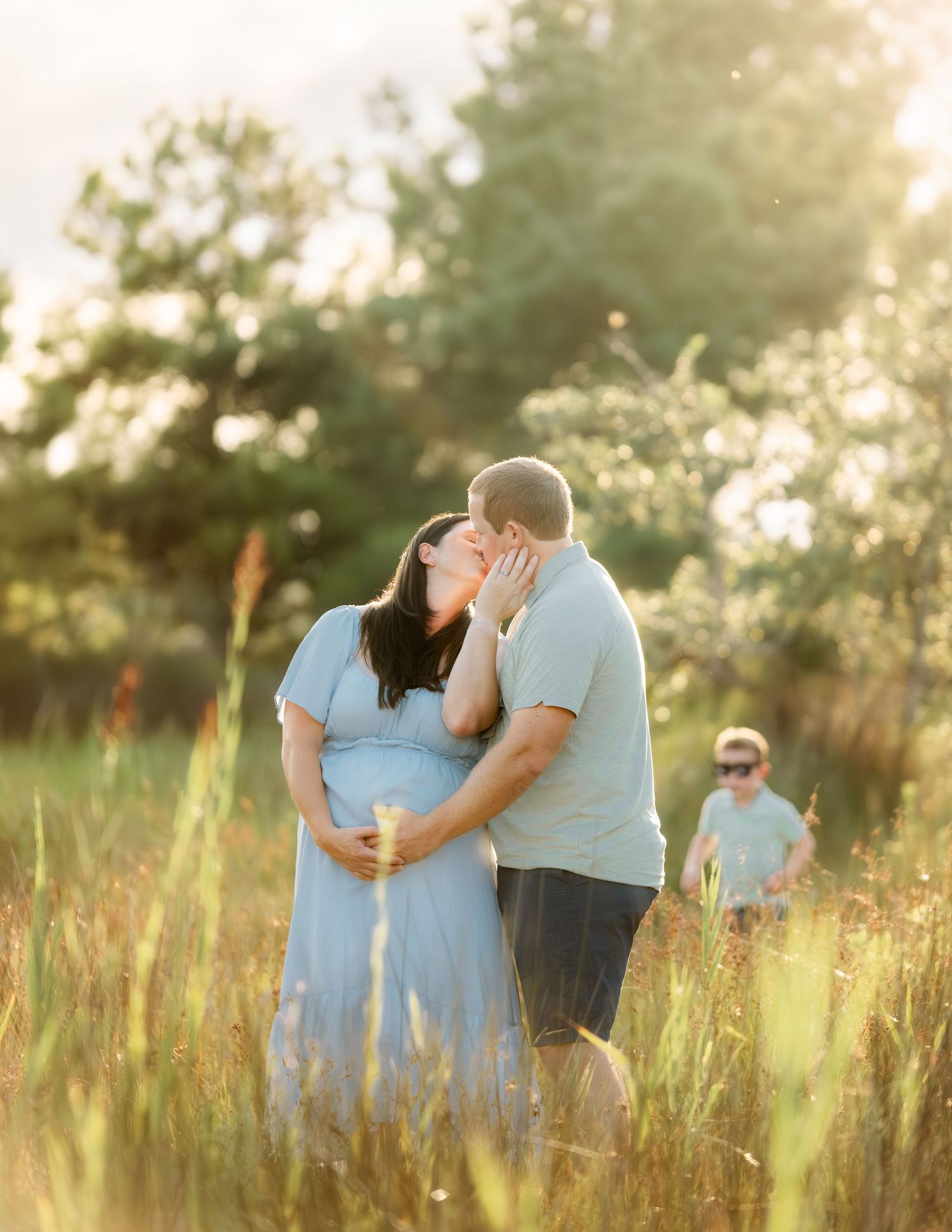 A romantic couple embraces in a sunlit field of tall grass during golden hour while someone watches in the background.
