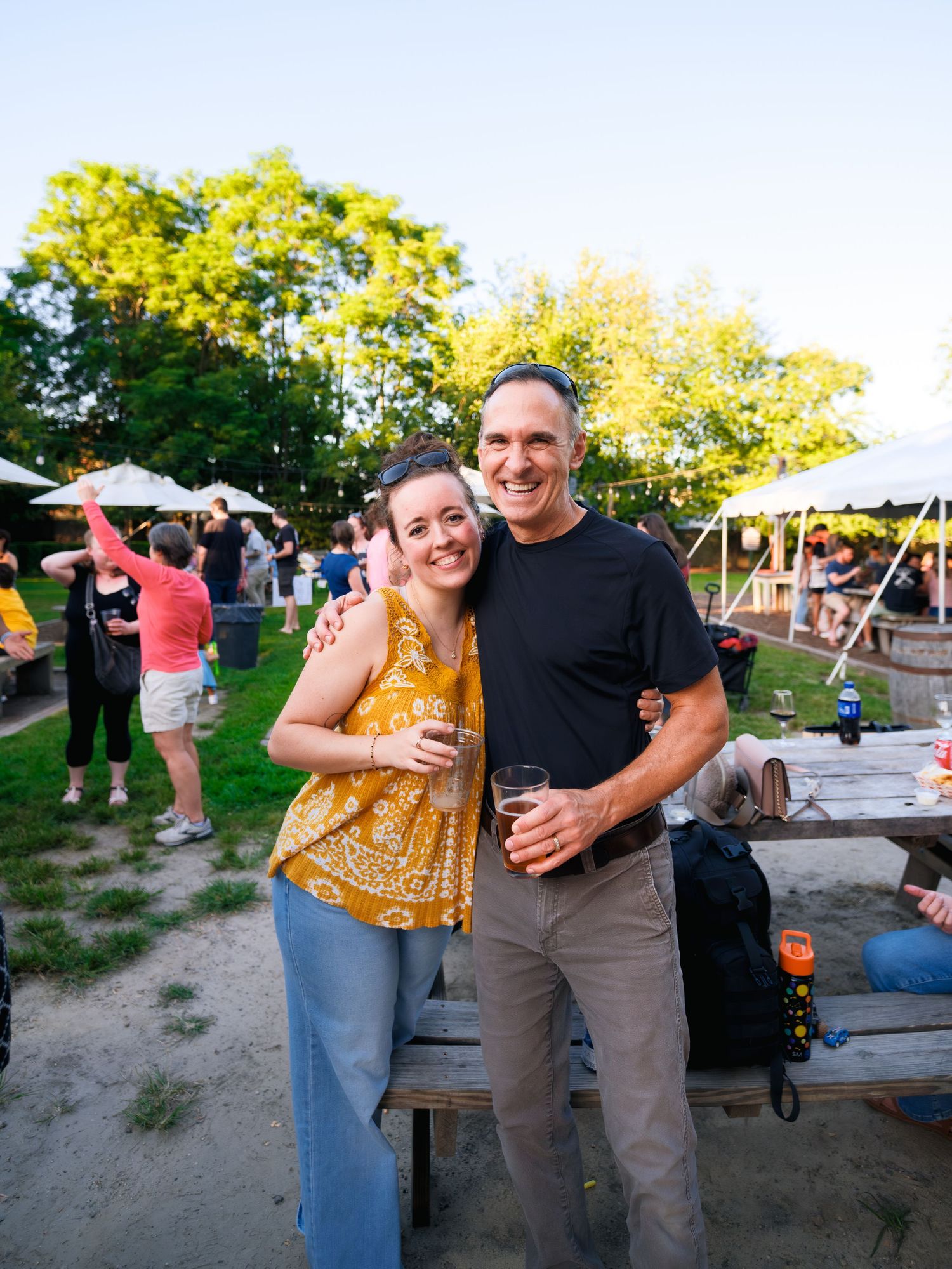 Guests smiling together at a casual outdoor event with white tents and trees visible in the background.