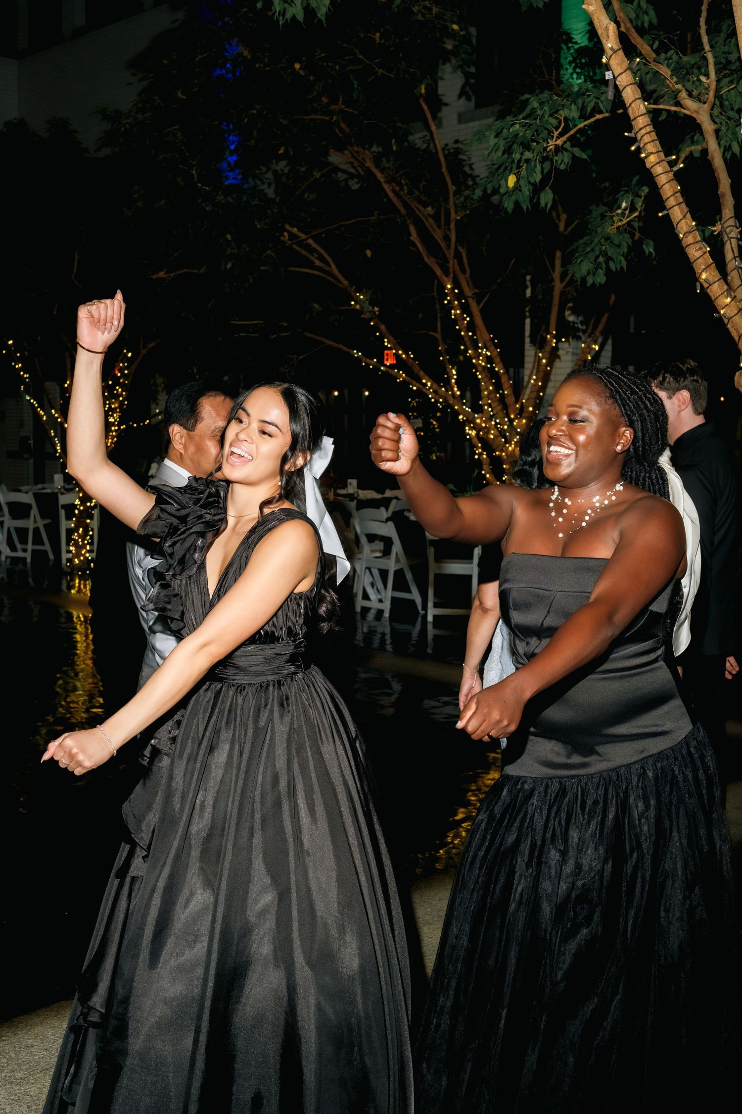 Two guests in elegant black formal gowns dancing joyfully at a nighttime event with twinkle-lit trees in background.