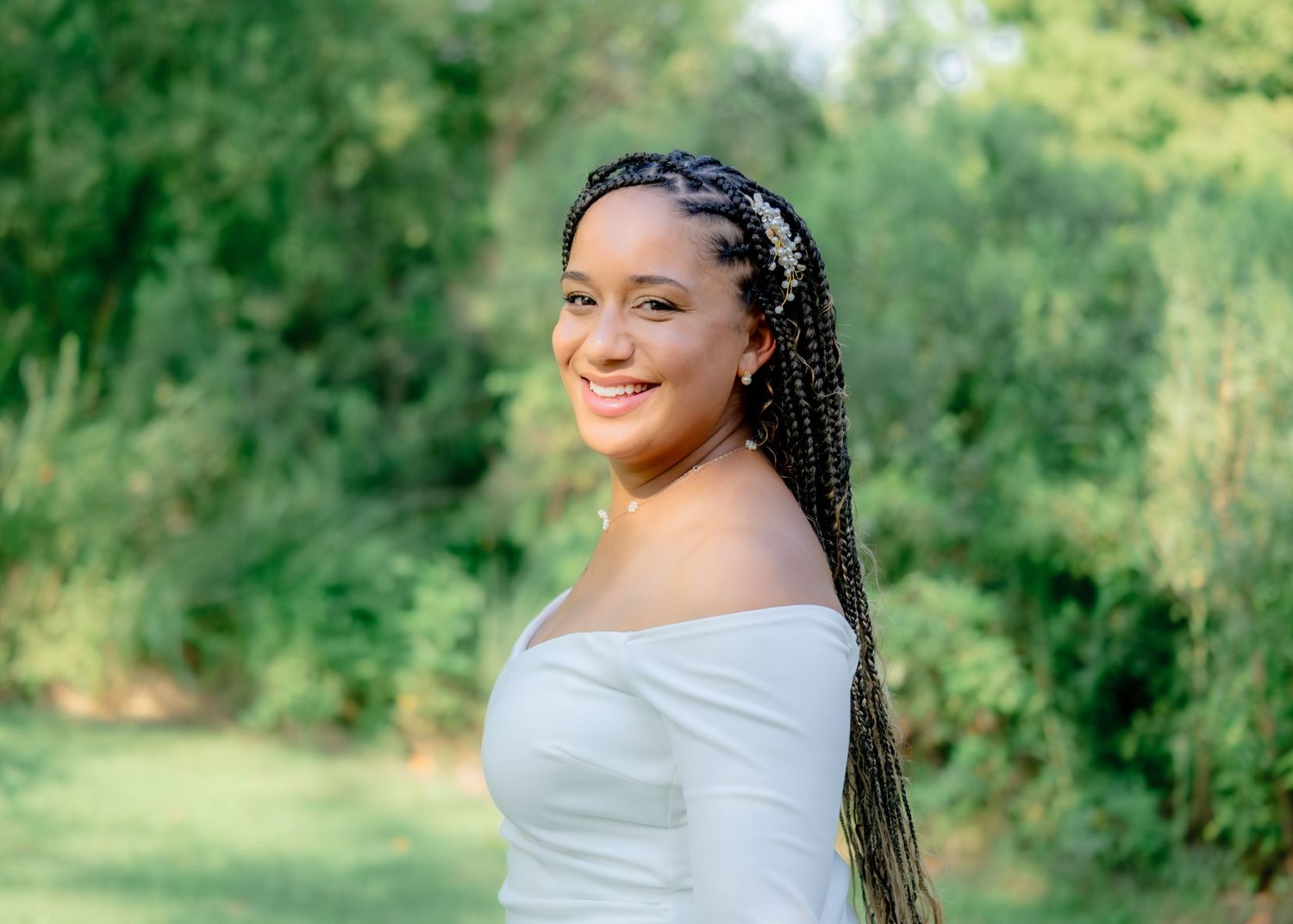 A person with braided hair poses outdoors in a white off-shoulder top amidst green foliage in a natural setting.