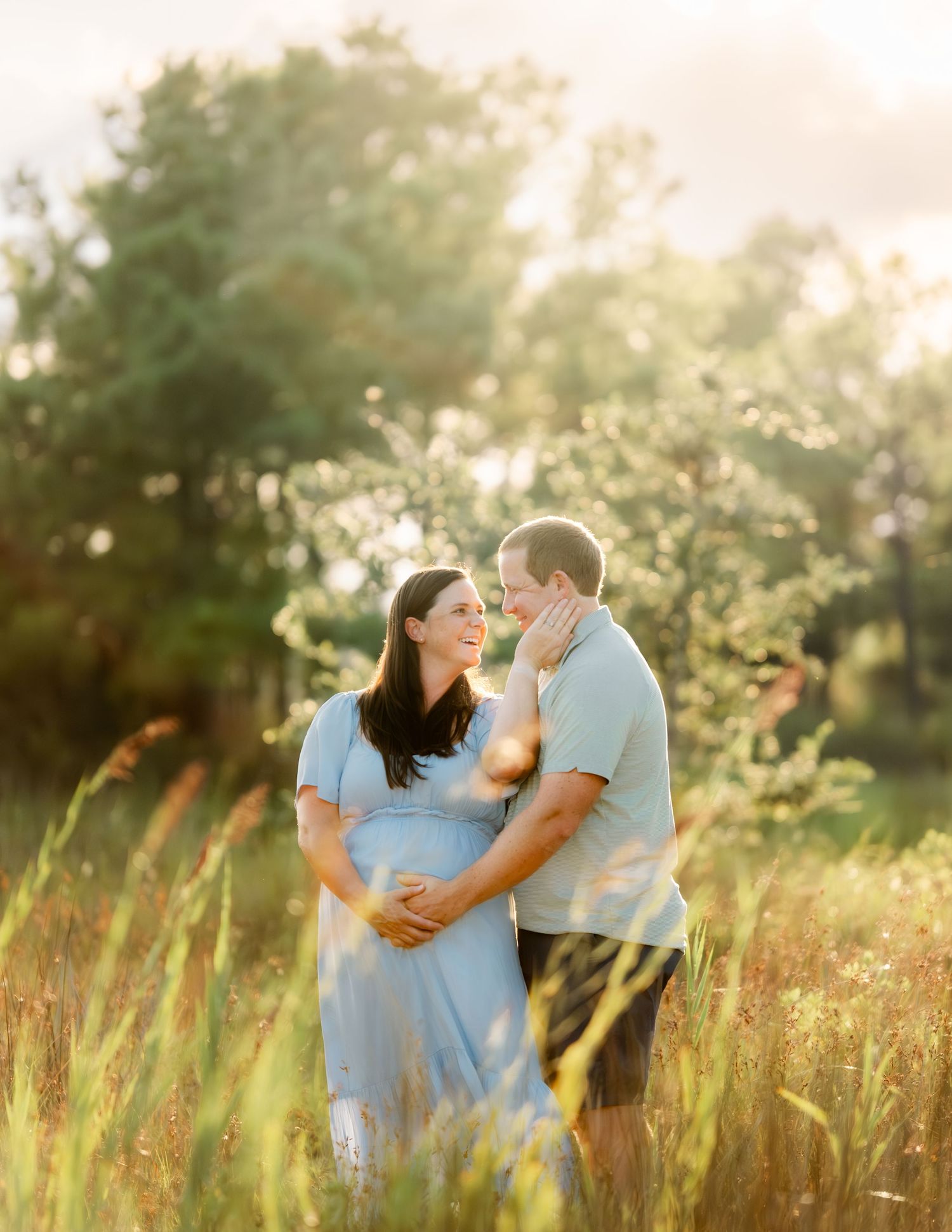 A romantic maternity photo shows a couple embracing in a sunlit field of tall grass during golden hour.