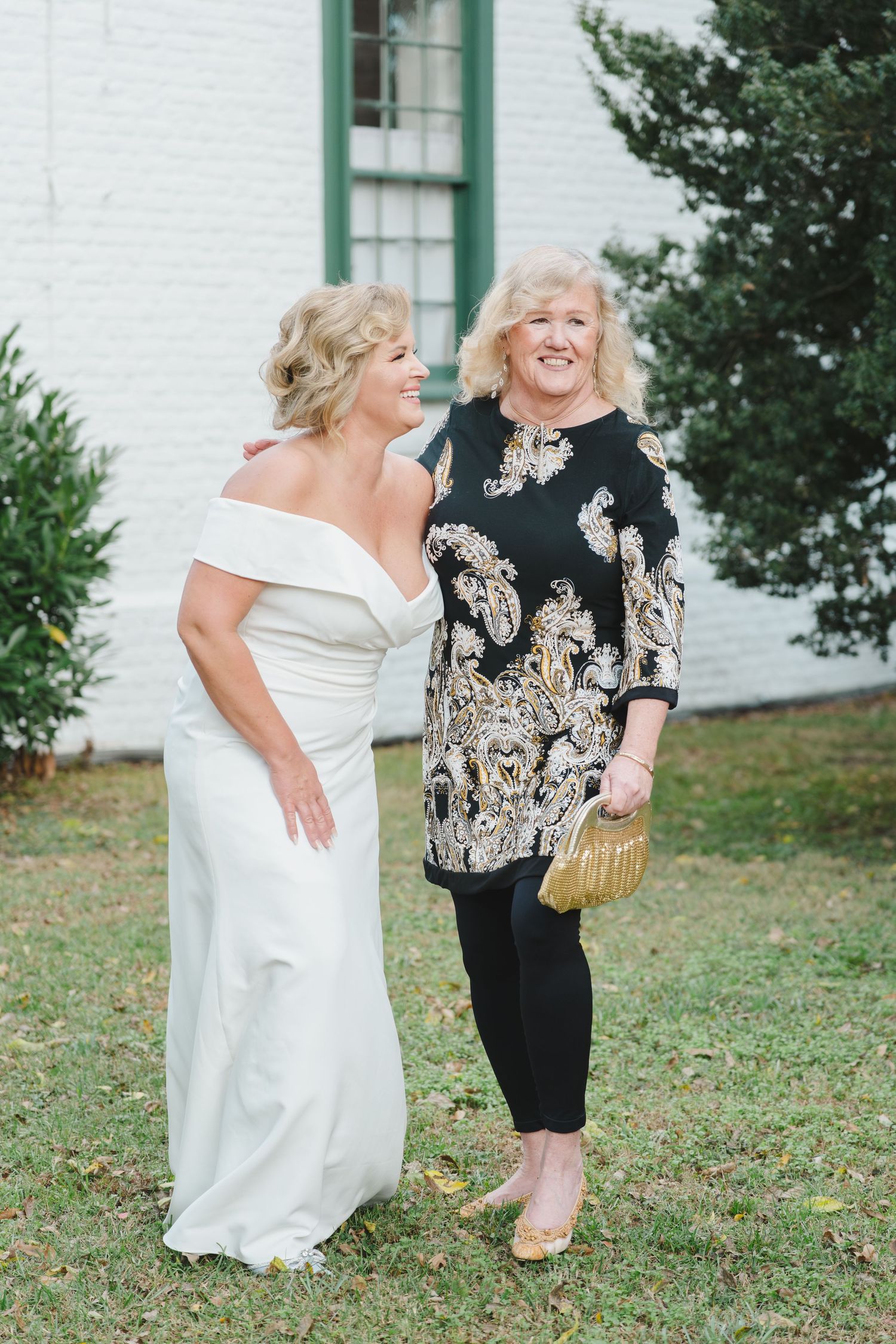 Two women share a joyful moment outdoors, one in an elegant white gown and another in a black and gold patterned dress.