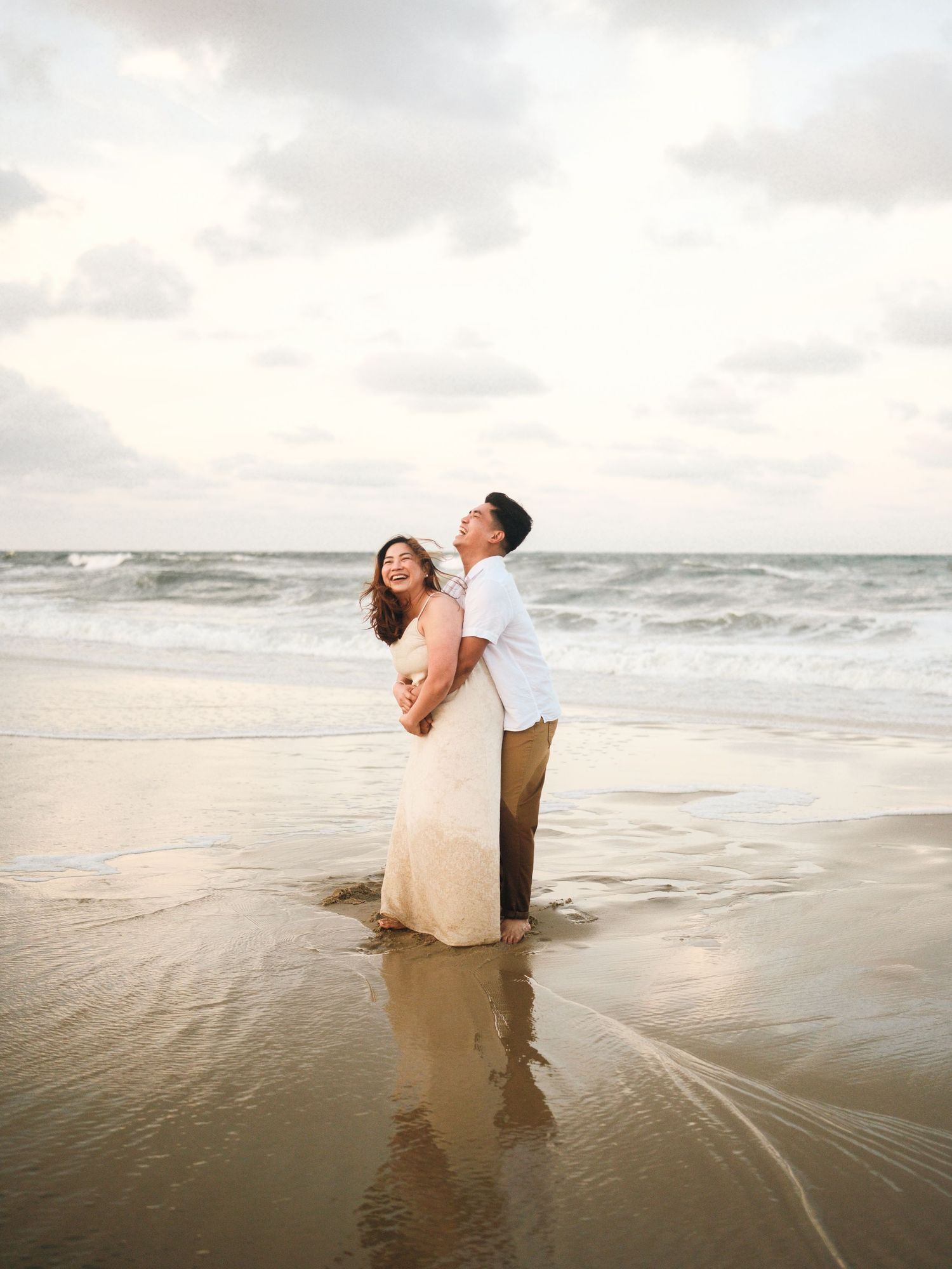 Playful couple dipping and dancing in ocean waves at beach during sunset.
