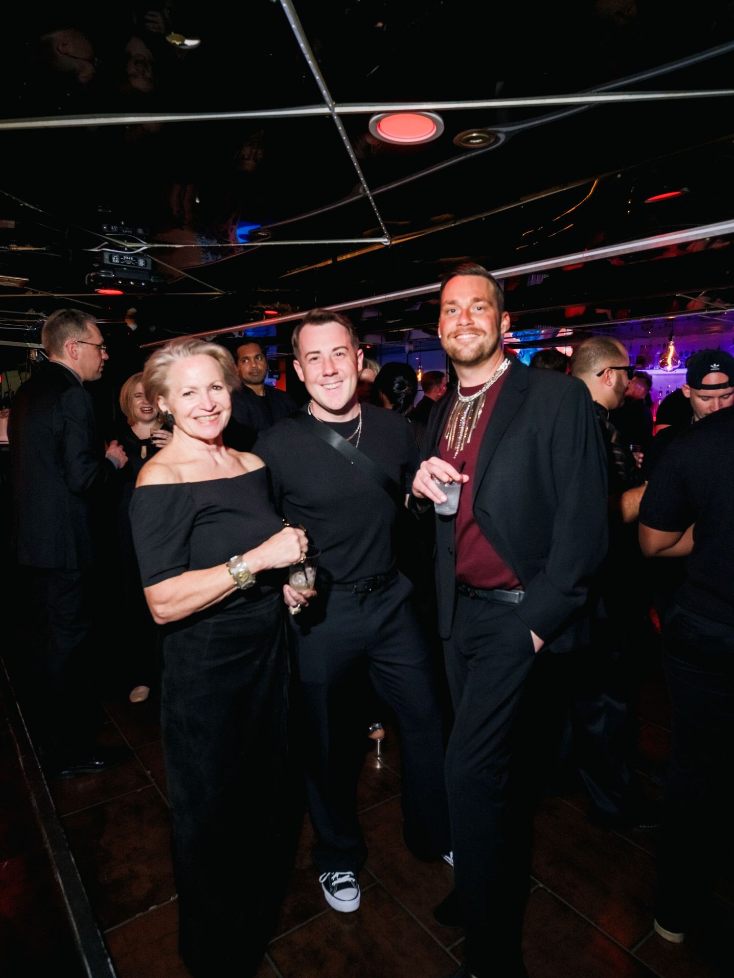 Three people pose together at a dimly lit nightclub with colorful lighting and exposed ceiling beams overhead.