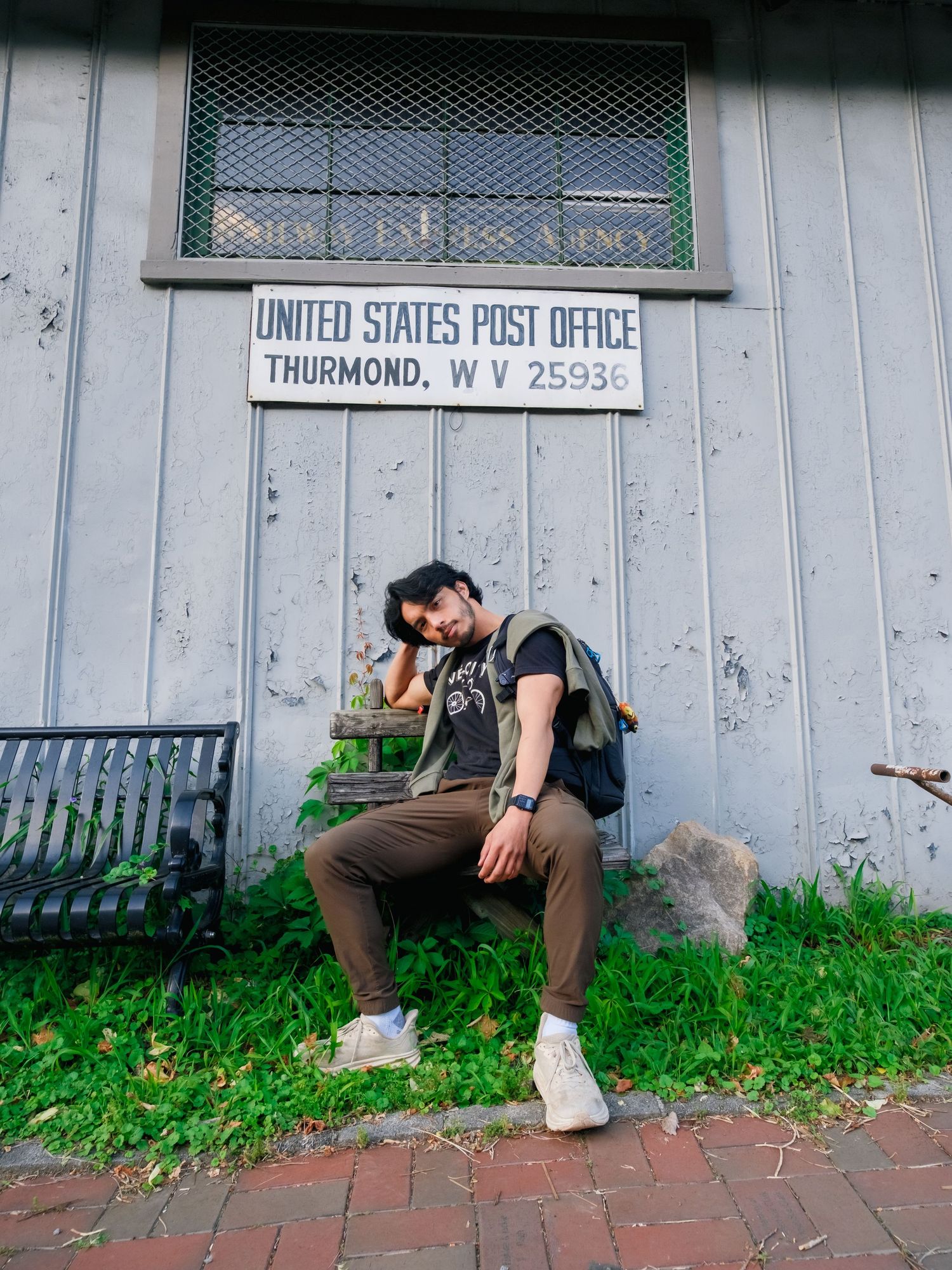 A traveler sits outside the United States Post Office in Thurmond, West Virginia, with a vintage brick pathway and green grass.