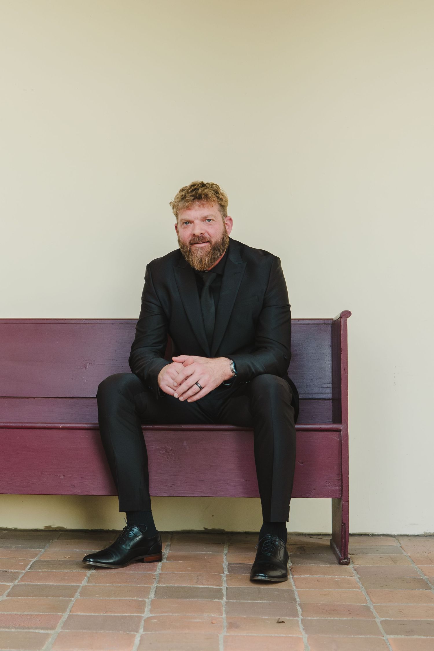 A person with a red beard wearing all black sits casually on a burgundy bench against a cream colored wall.