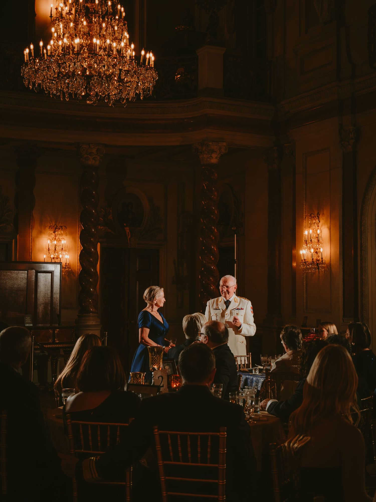 Wedding guests gathered in an elegant ballroom with ornate chandeliers and warm lighting during an evening reception.