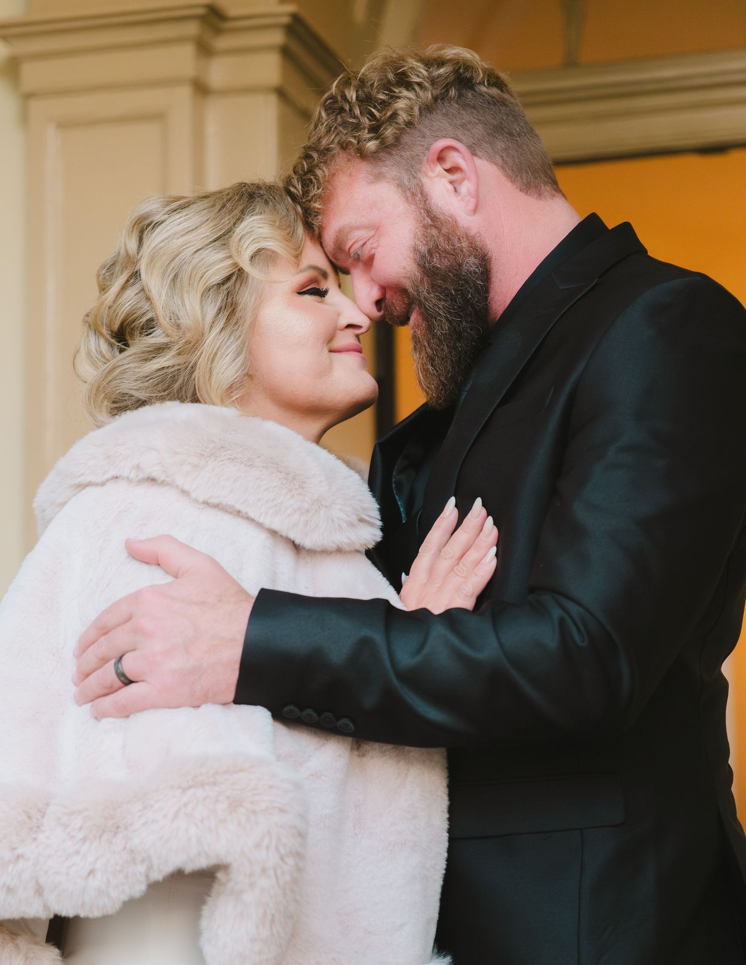 A couple shares an intimate moment in formal attire, with the woman wearing a white fur wrap and the man in a black suit.