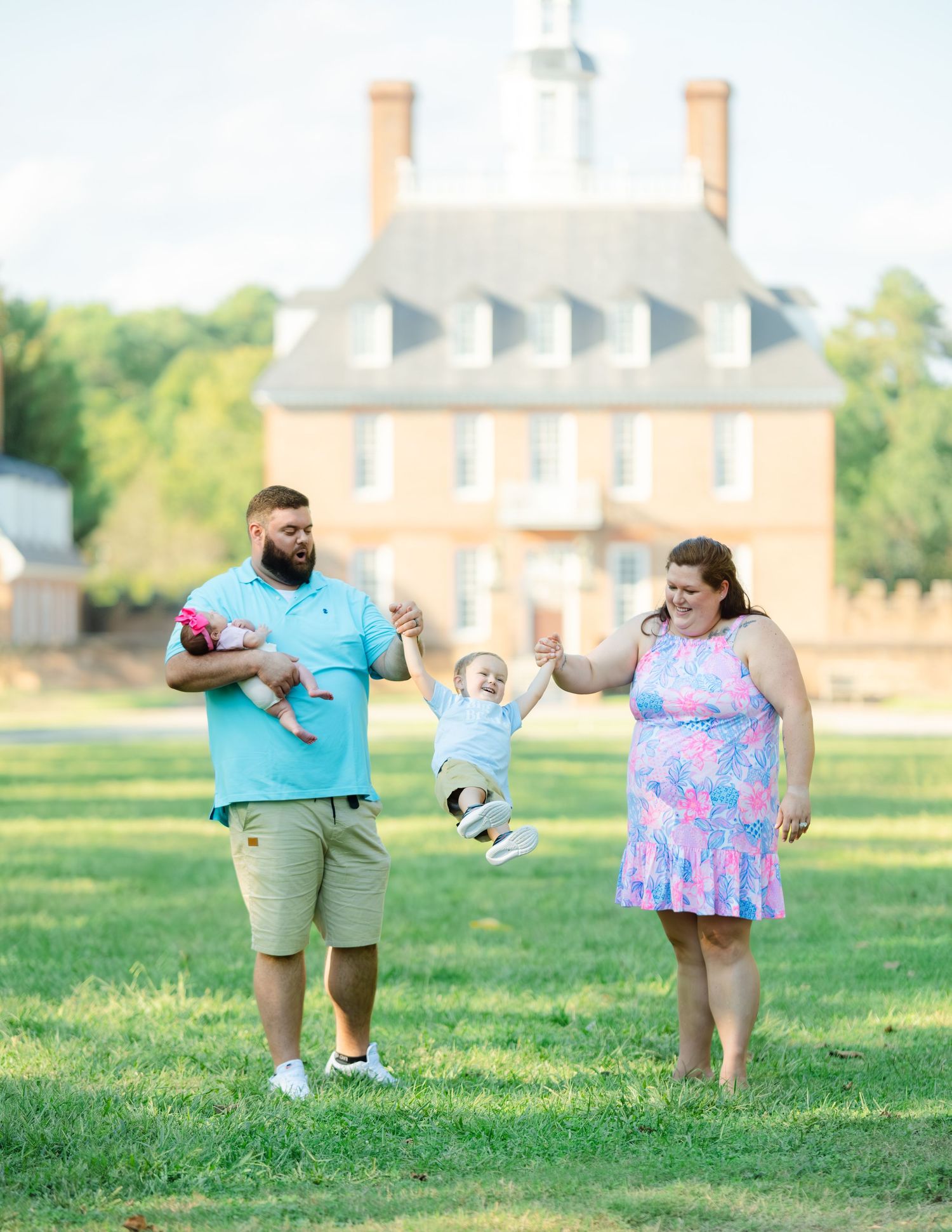 Family enjoying a playful moment together on a sunny green lawn with colonial buildings in the background.