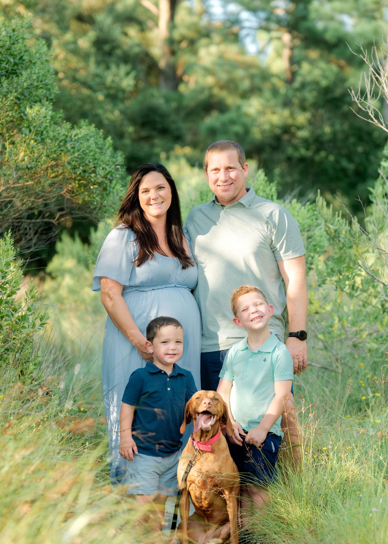 A family poses outdoors with their golden Vizsla dog on a sunny day among green foliage.
