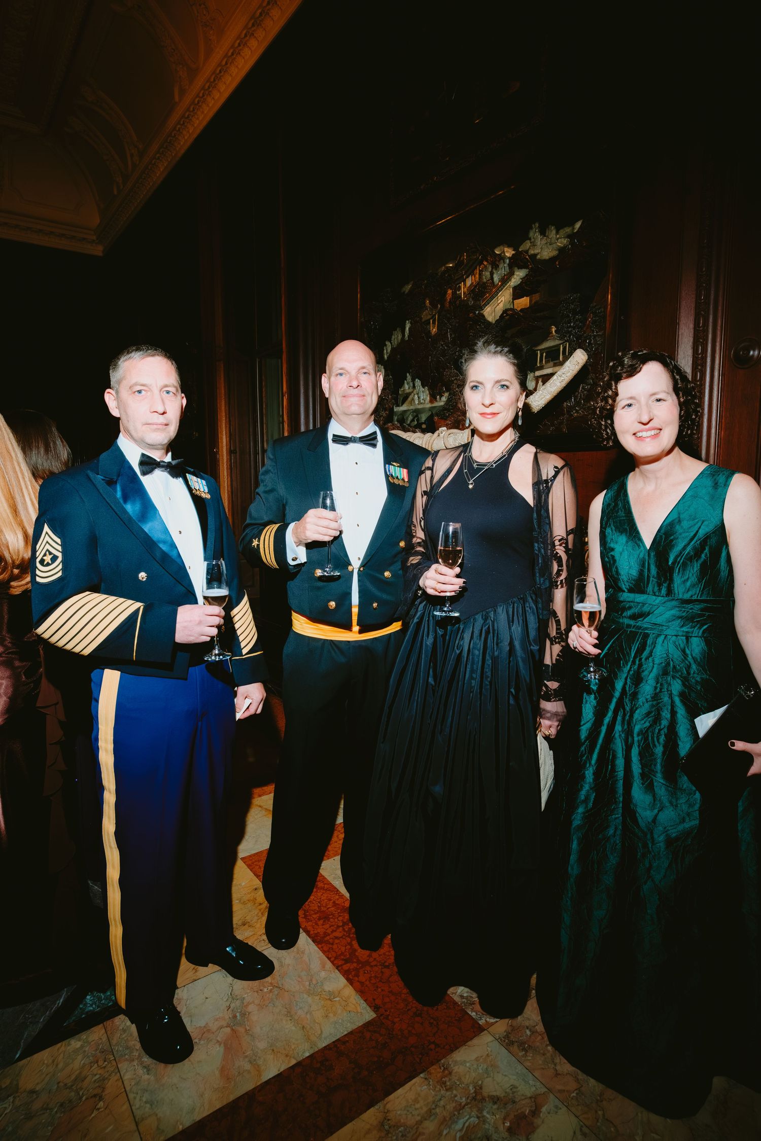 Guests in formal military dress uniforms and elegant evening gowns attend a glamorous formal event in an ornate room.