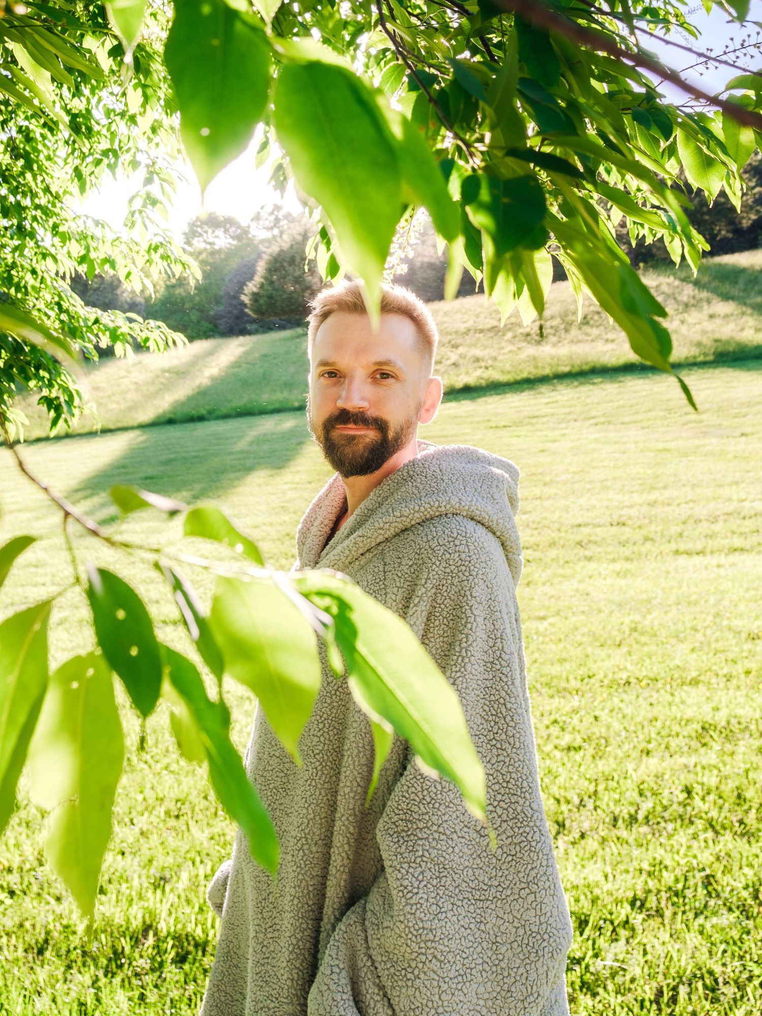 Person wearing a cozy gray fleece blanket stands in front of sunlit green leaves and grass on a summer morning.