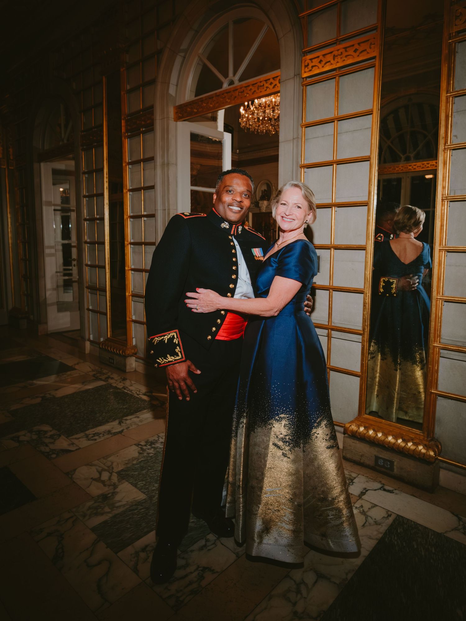 A couple in formal attire poses together at an elegant event with mirrored walls and chandeliers in the background.