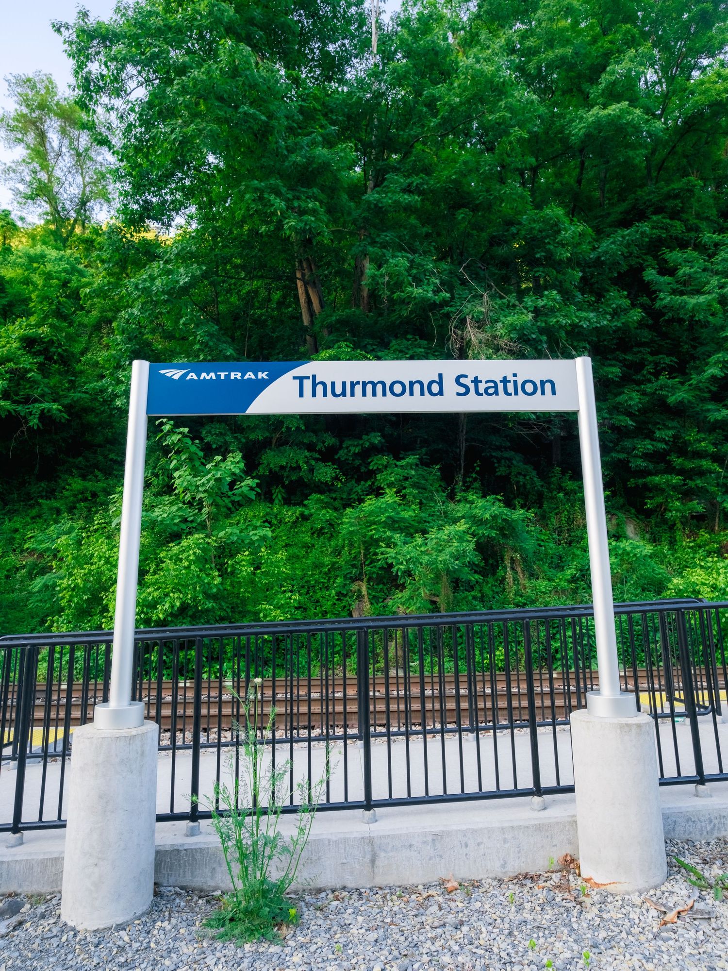 Amtrak station sign for Thurmond Station mounted on concrete pillars with black metal railings and green forest backdrop.