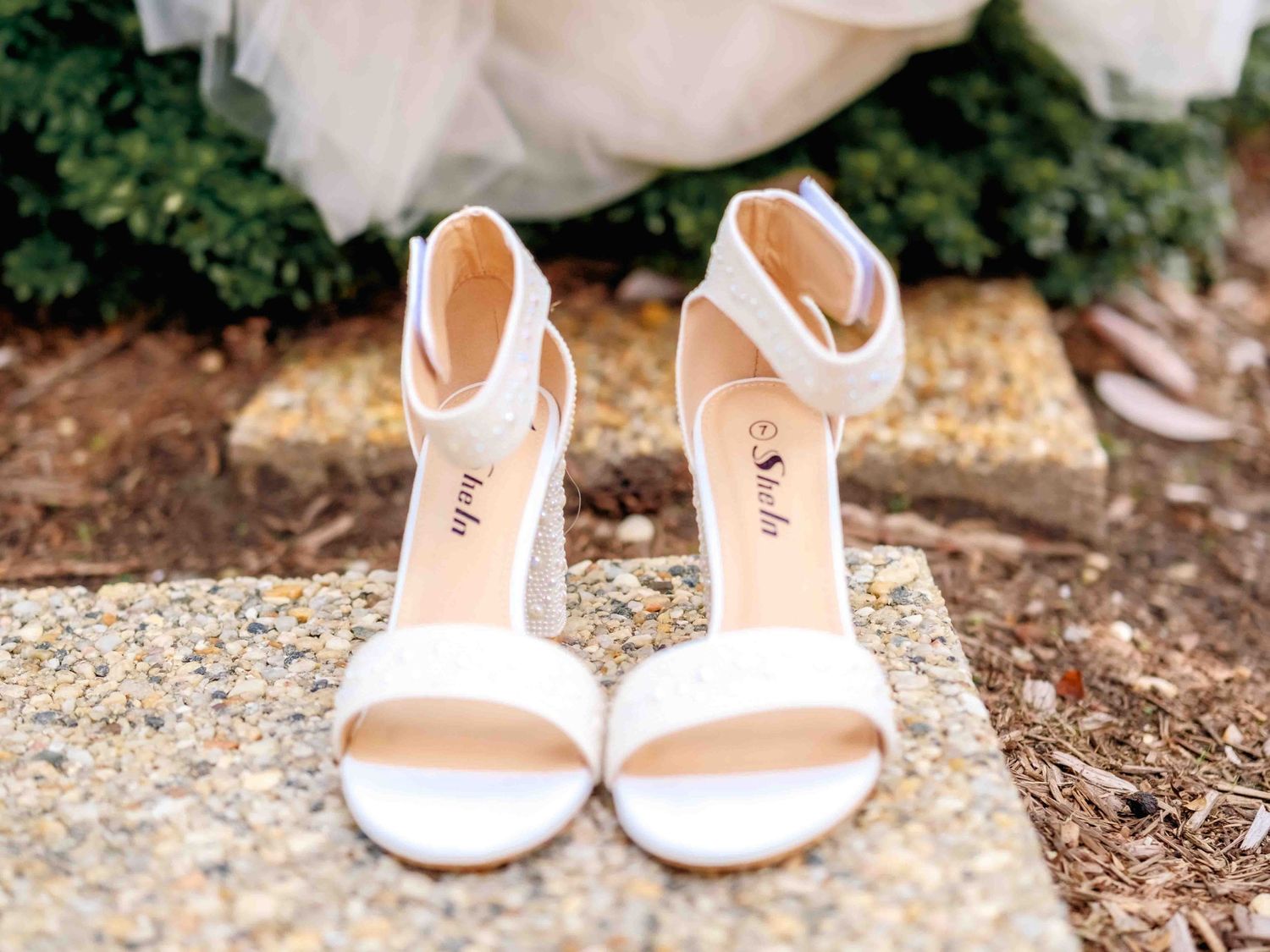 Elegant white bridal sandals with ankle straps sit on a stone surface surrounded by flowers.