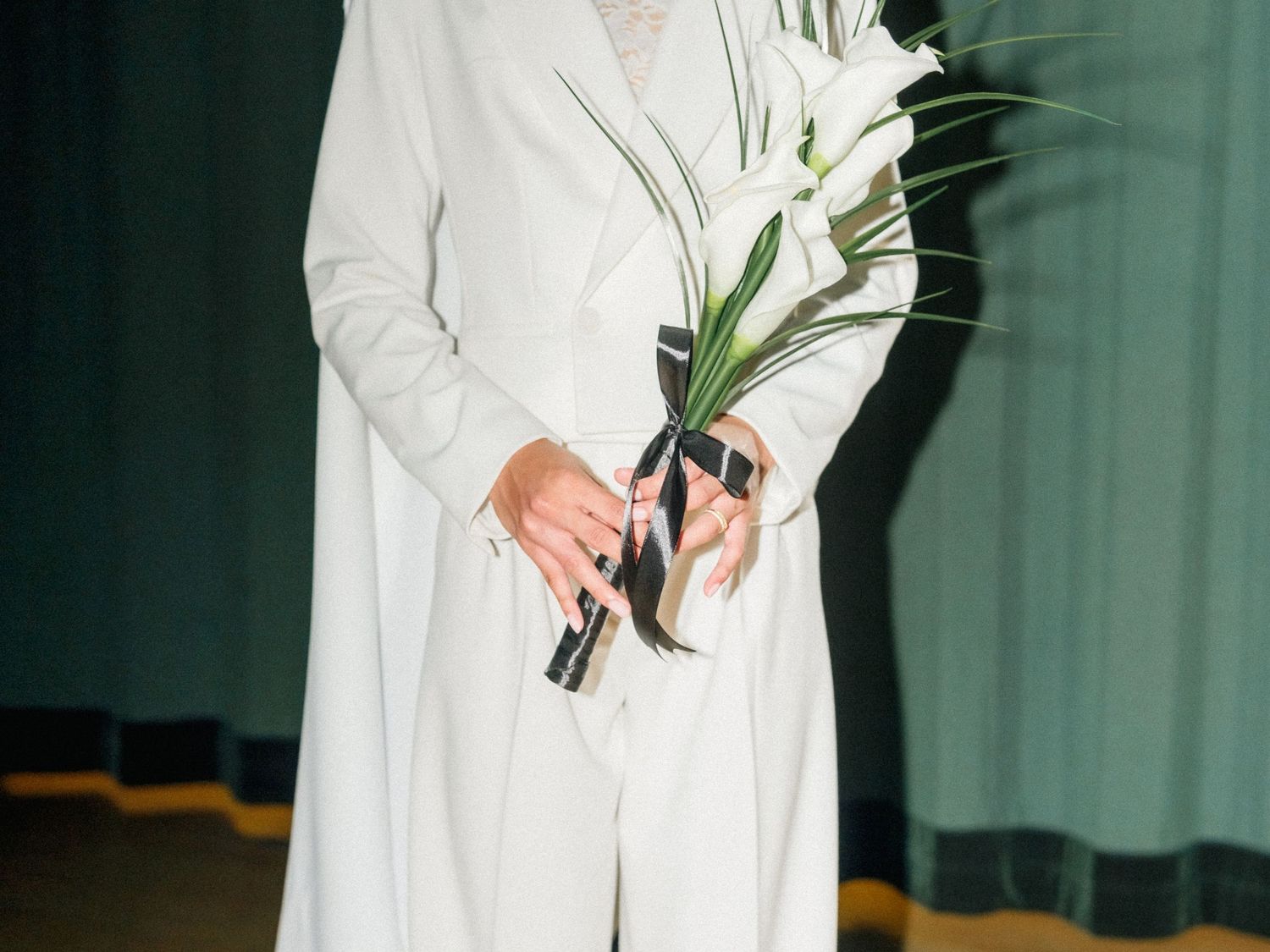 Someone in a white suit holds a single white lily flower against a green curtain backdrop.