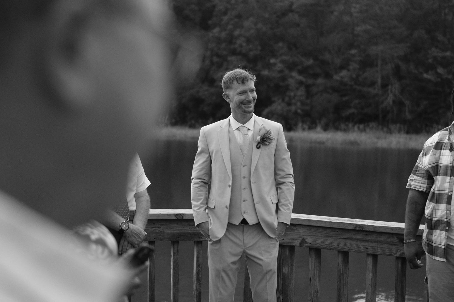 Black and white wedding photo of someone in a light colored suit smiling while standing on a wooden dock near a lake.