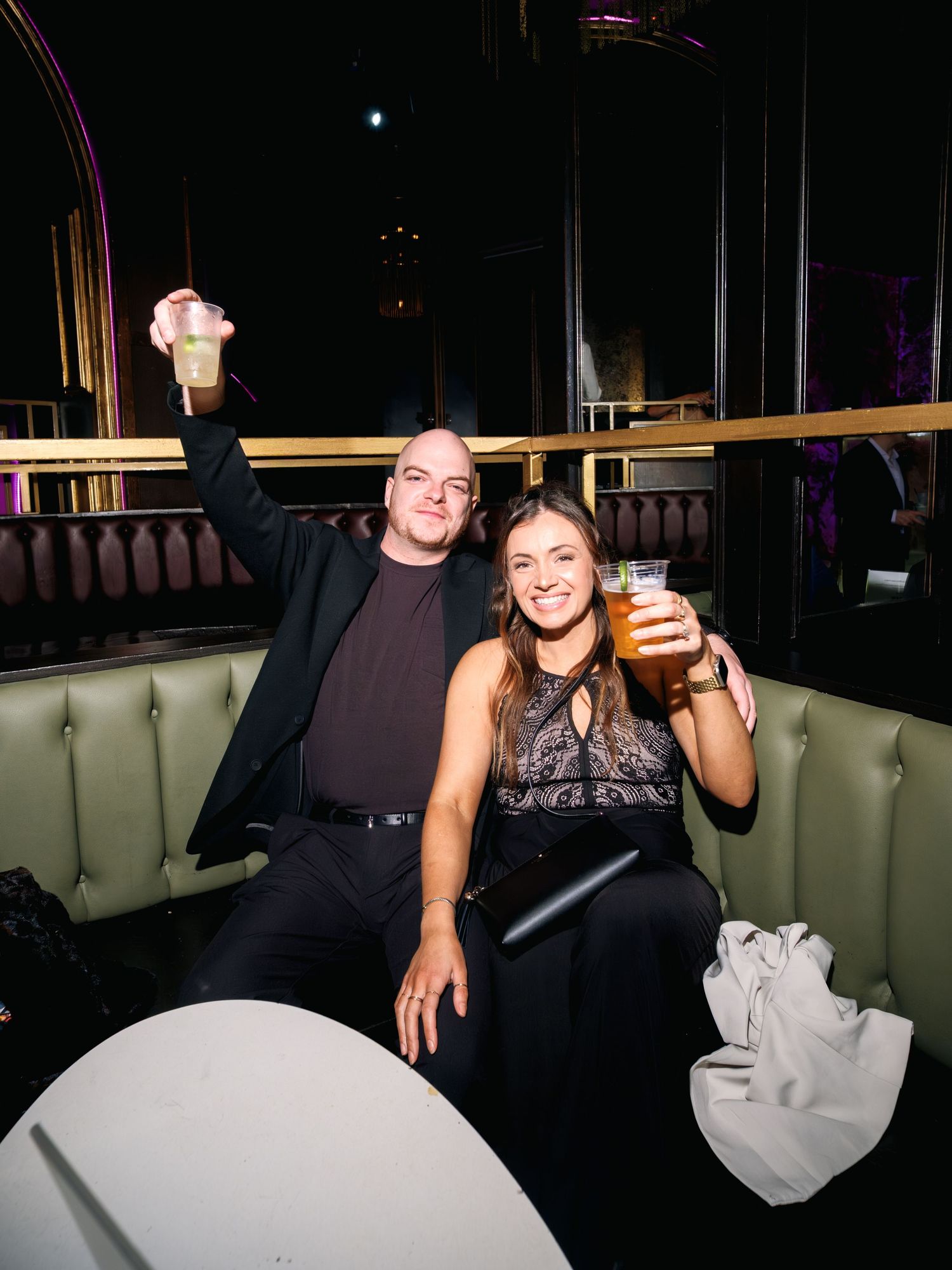 Two people sit in a dimly lit lounge booth raising their glasses in a celebratory toast.