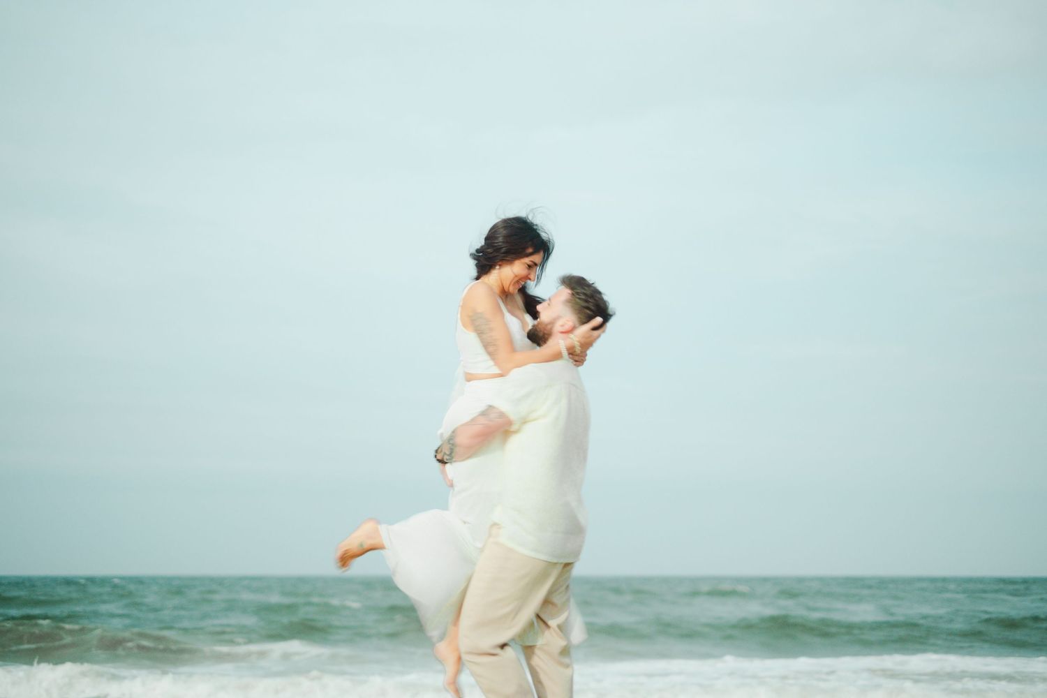 A romantic couple in white outfits embraces on a beach with ocean waves and cloudy skies in the background.
