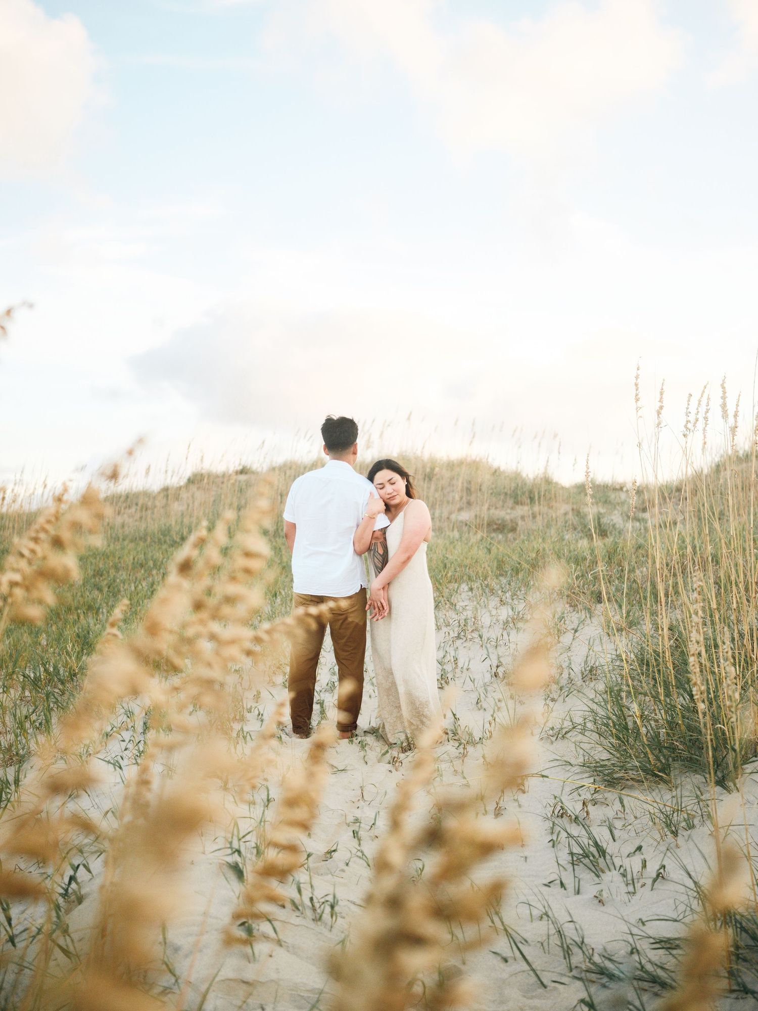 Couple walking through coastal dunes with tall seagrass at sunset.