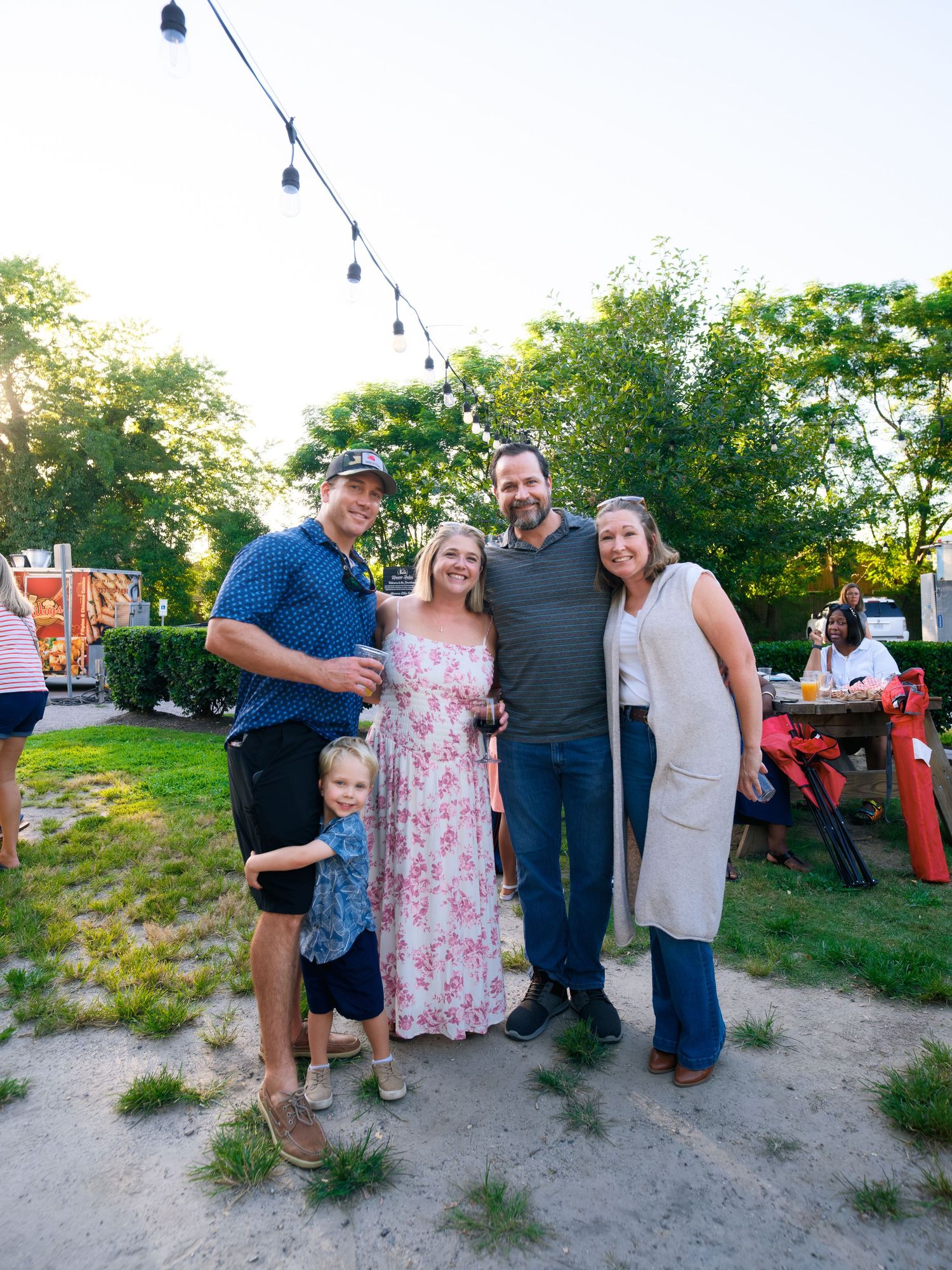 People enjoying an outdoor gathering on sandy ground with string lights and trees in the background.