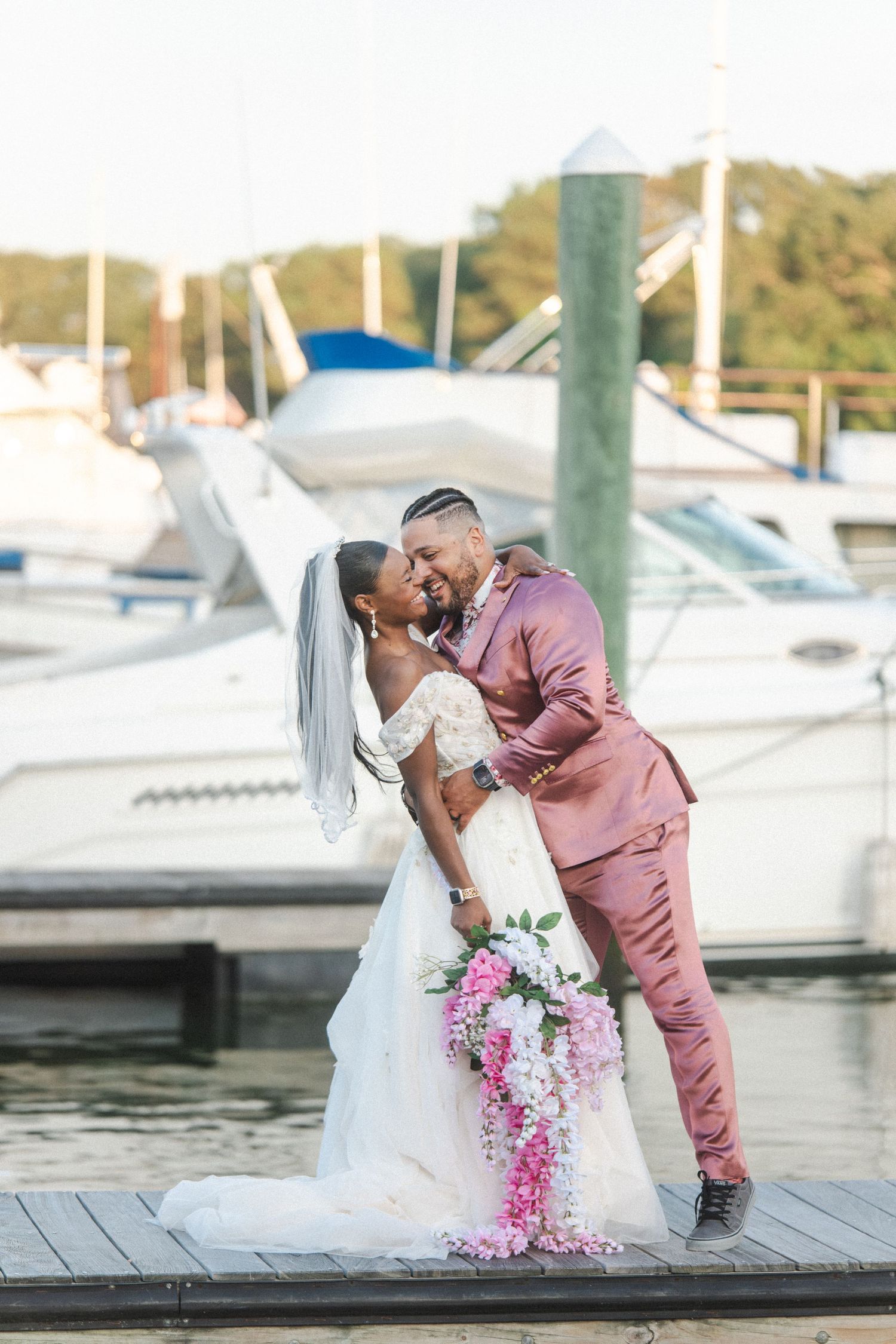 A romantic wedding couple shares a tender moment on a marina dock surrounded by boats on a sunny day.
