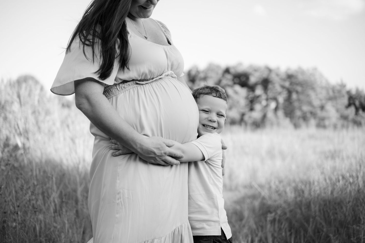 Black and white maternity photo series showing a pregnant woman and child embracing in a field at sunset.