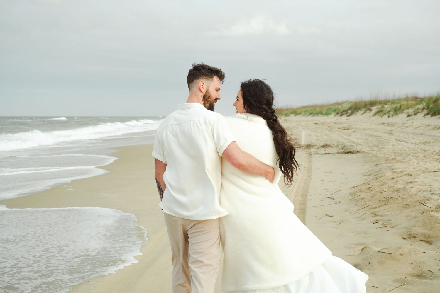 A couple in wedding attire stands together on a peaceful sandy beach along the ocean shore.