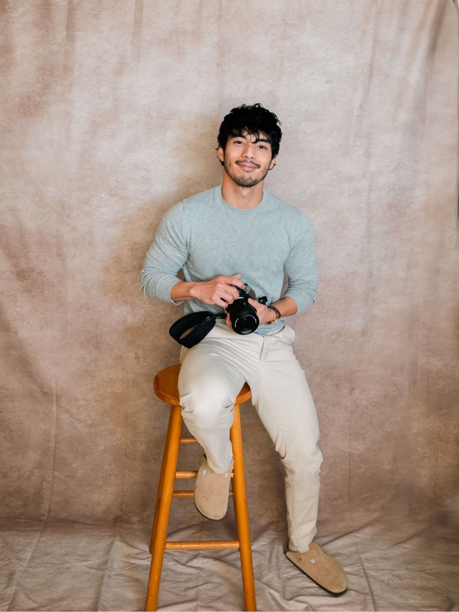 Photographer in casual grey sweater and khaki pants sits on wooden stool holding camera in studio setting.