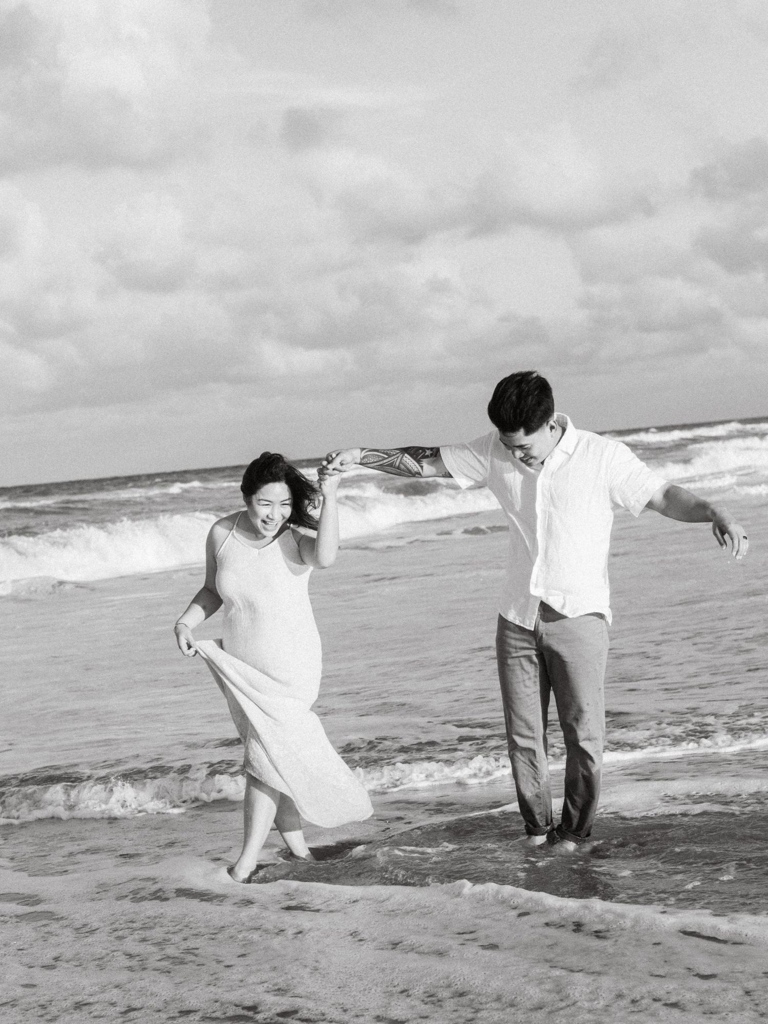 A couple in casual attire dances playfully along the beach shoreline in black and white photography.