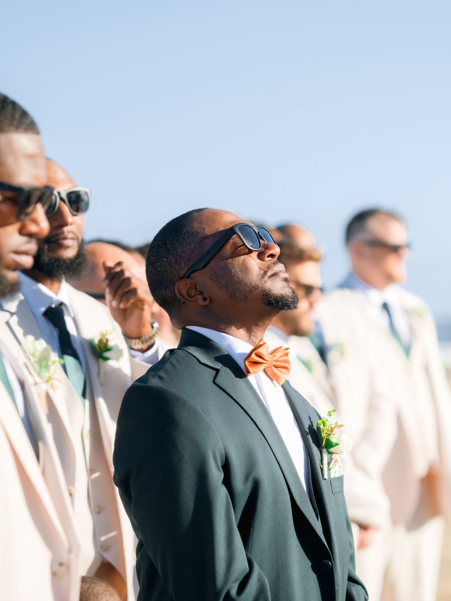 Wedding party members in sunglasses and formal attire pose against blue sky.
