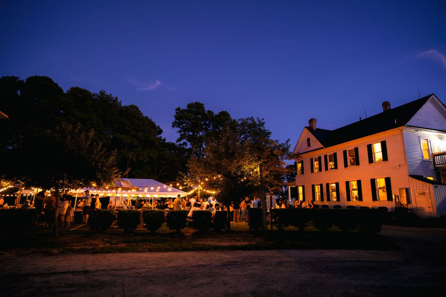 A rustic nighttime outdoor wedding reception with string lights illuminating a white colonial house and garden setting.