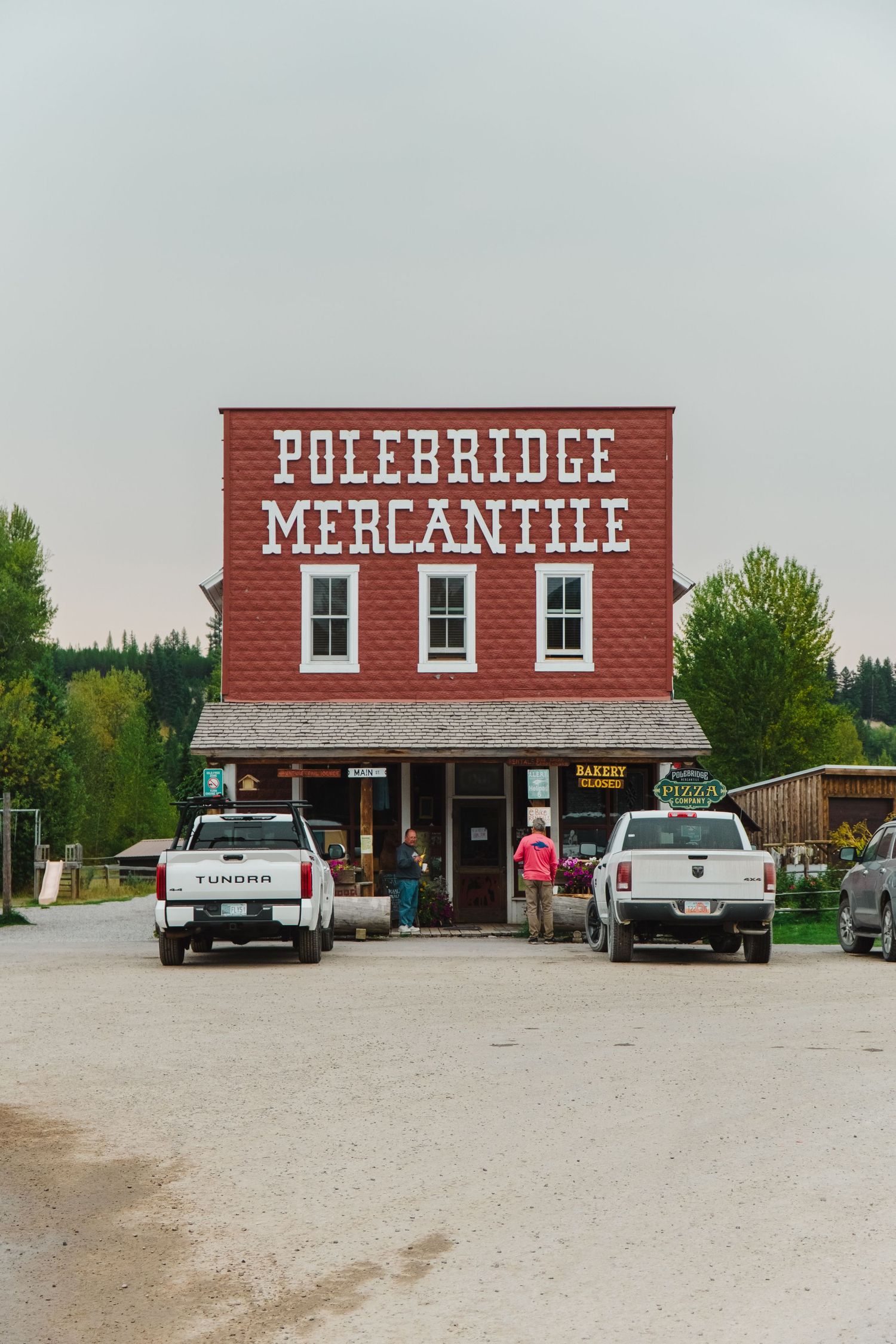 View of Polebridge Mercantile, a historic red building with white trim and vintage cars parked outside in rural Montana.