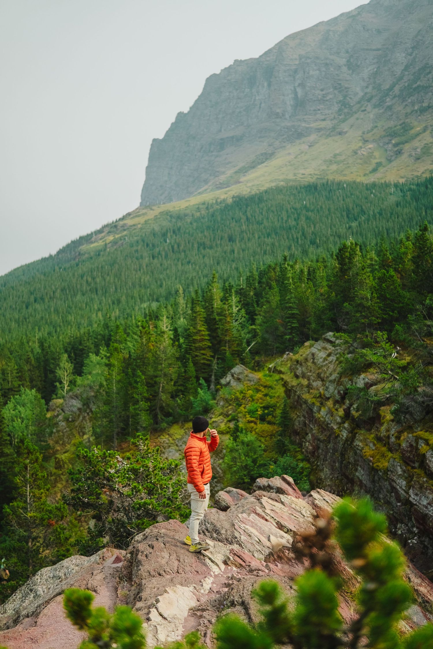 Hiker in orange jacket stands on rocky outcrop overlooking scenic mountain valley with evergreen forest in Glacier National Park.