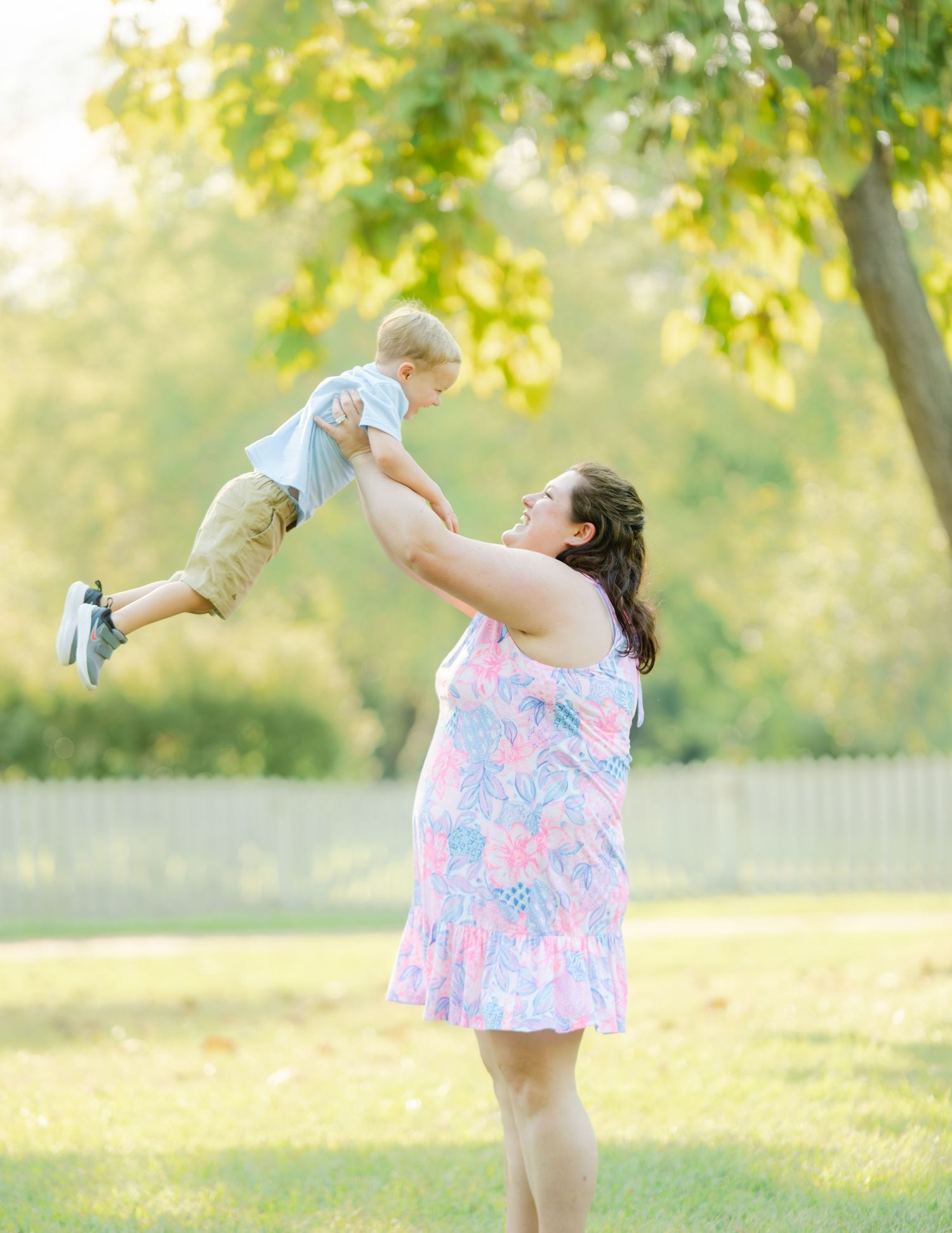 A joyful moment captured in a sunlit park as a woman in a floral dress lifts a small child up in the air.