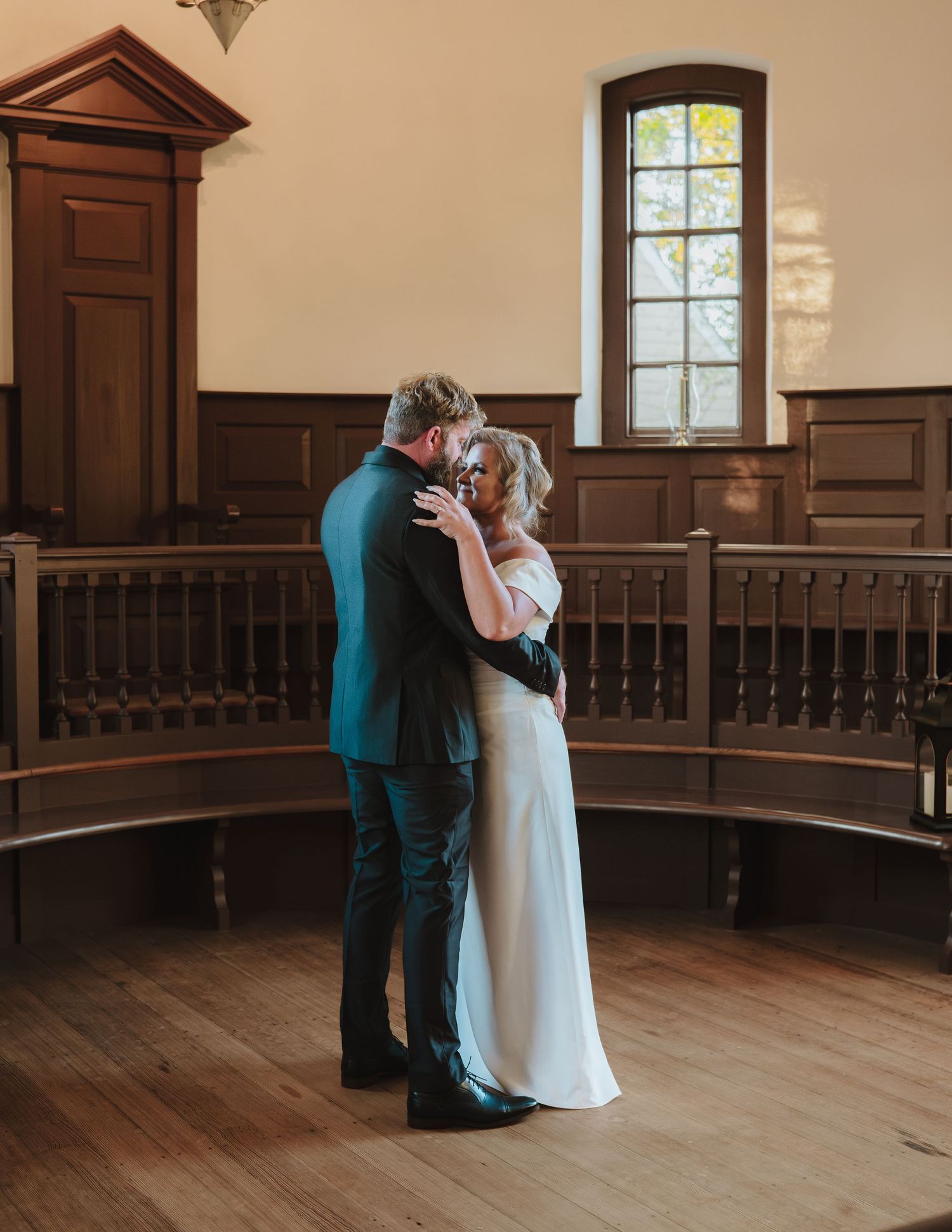 Two people share an intimate moment while standing in a historic hall with dark wood paneling and an arched window.