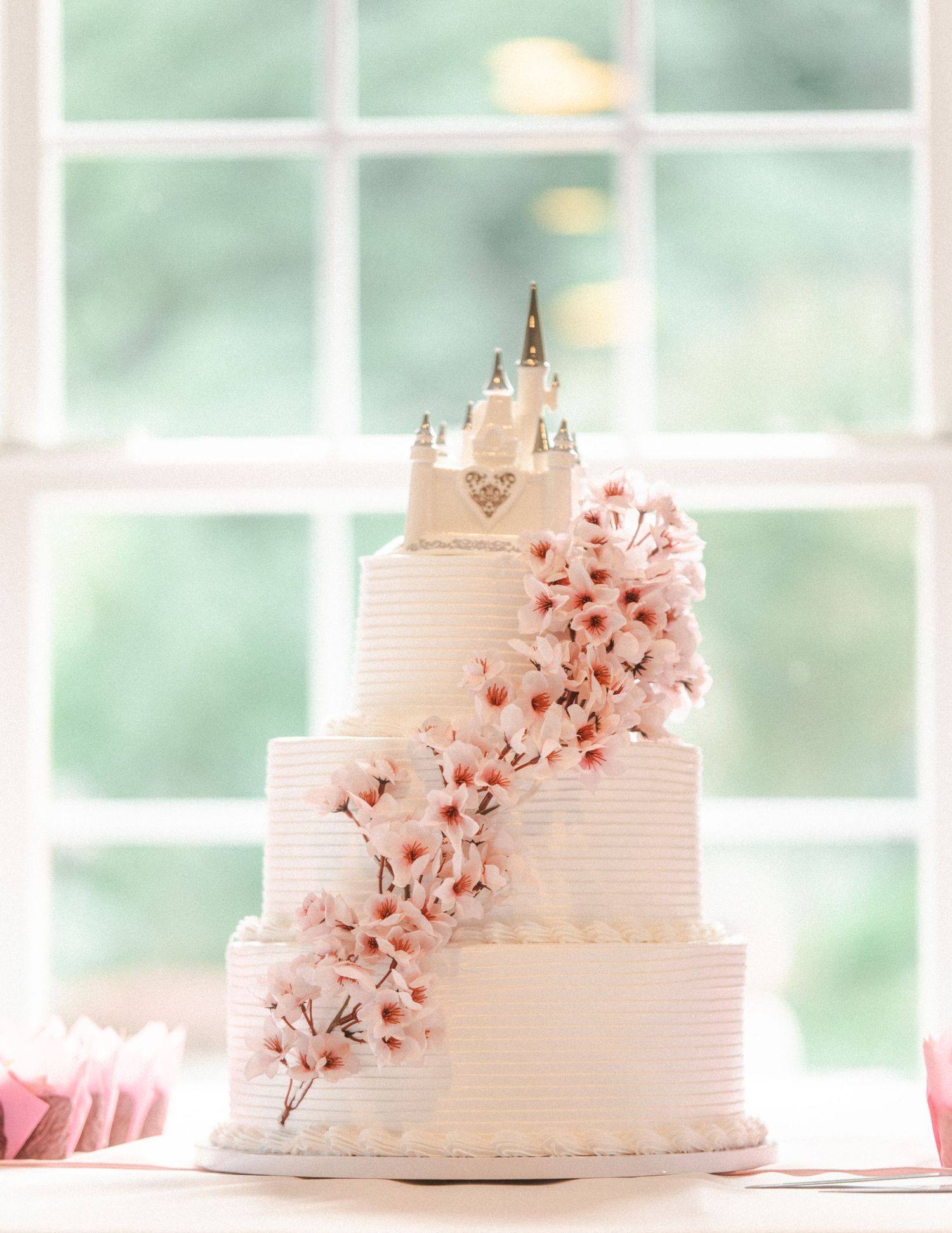 Elegant three-tier white wedding cake with pink floral accents and castle topper displayed in front of window.