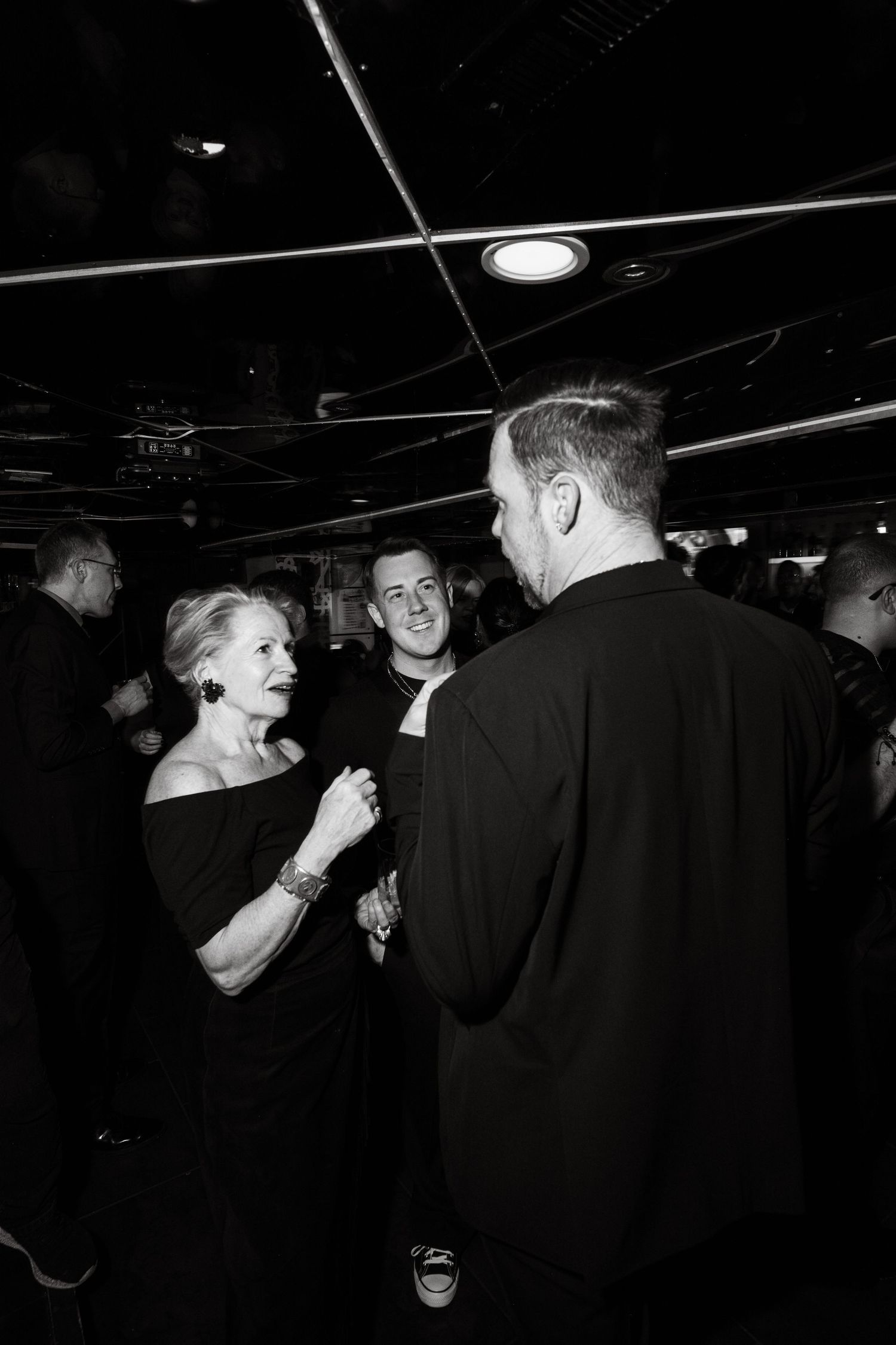 A black and white photo of people engaged in animated conversation at a dark venue with overhead lighting.