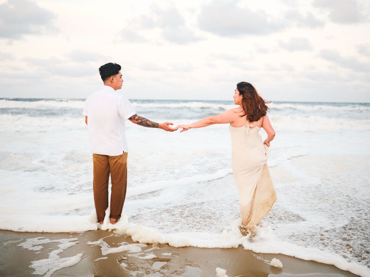 A playful couple in white and tan clothing dance and twirl together by the ocean waves.