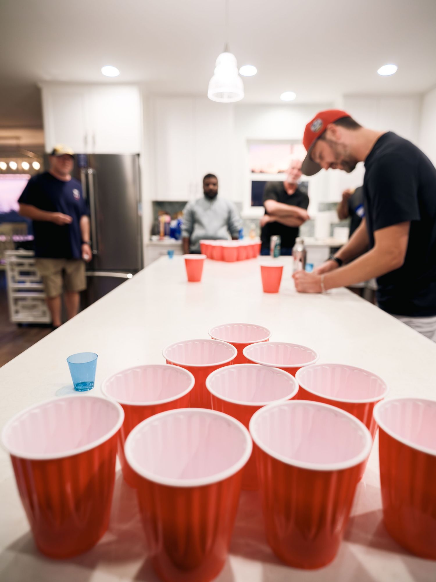 Red plastic cups arranged for beer pong game on white countertop with people standing in kitchen background.
