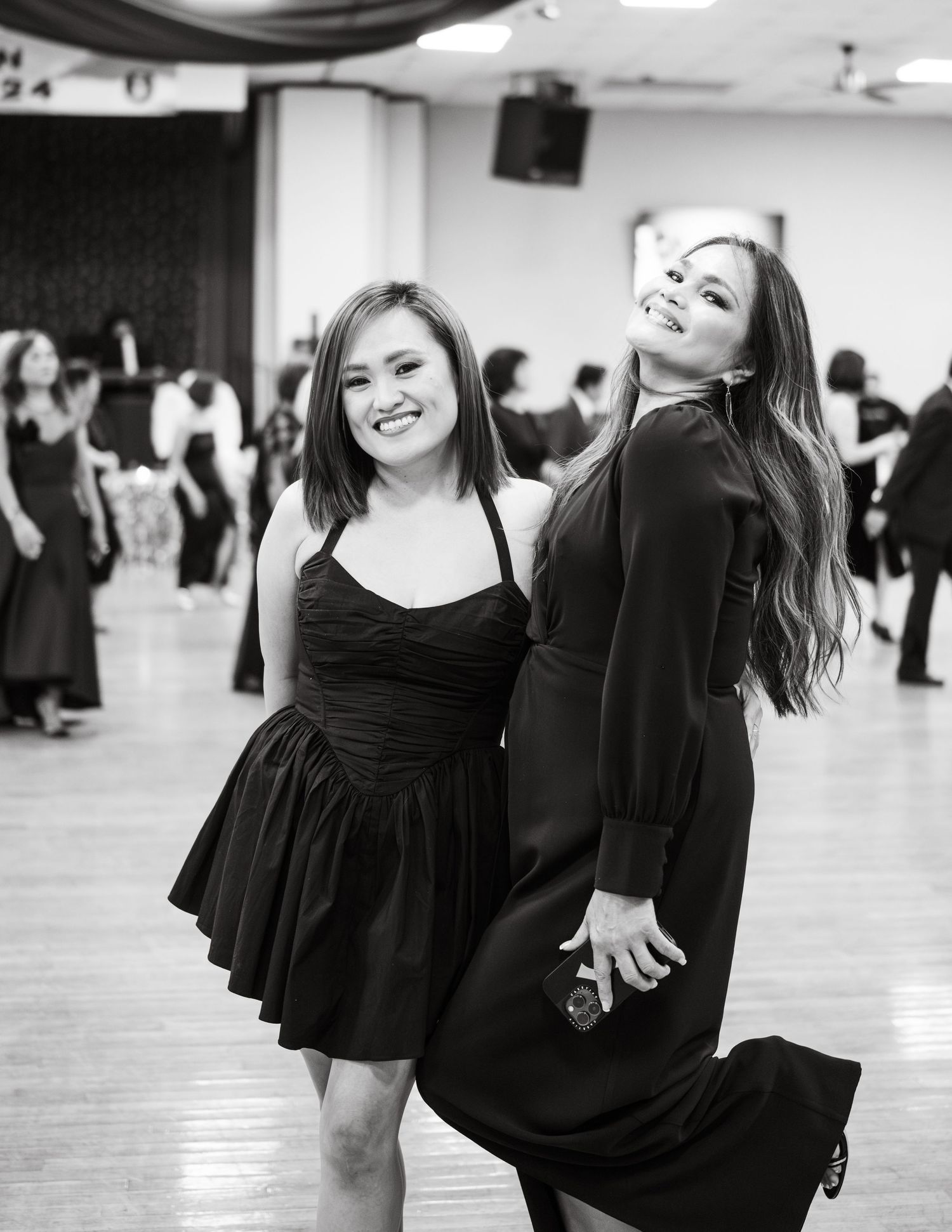 Two dancers in black dresses strike playful poses on a dance floor with other dancers visible in the background.