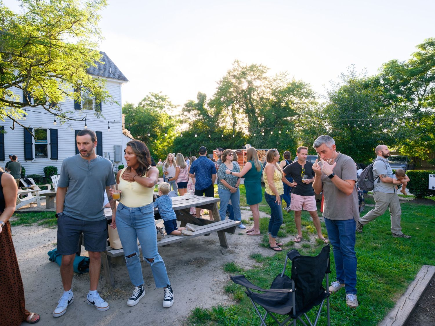 A casual outdoor social gathering with string lights hanging above as guests mingle on a lawn near a house.