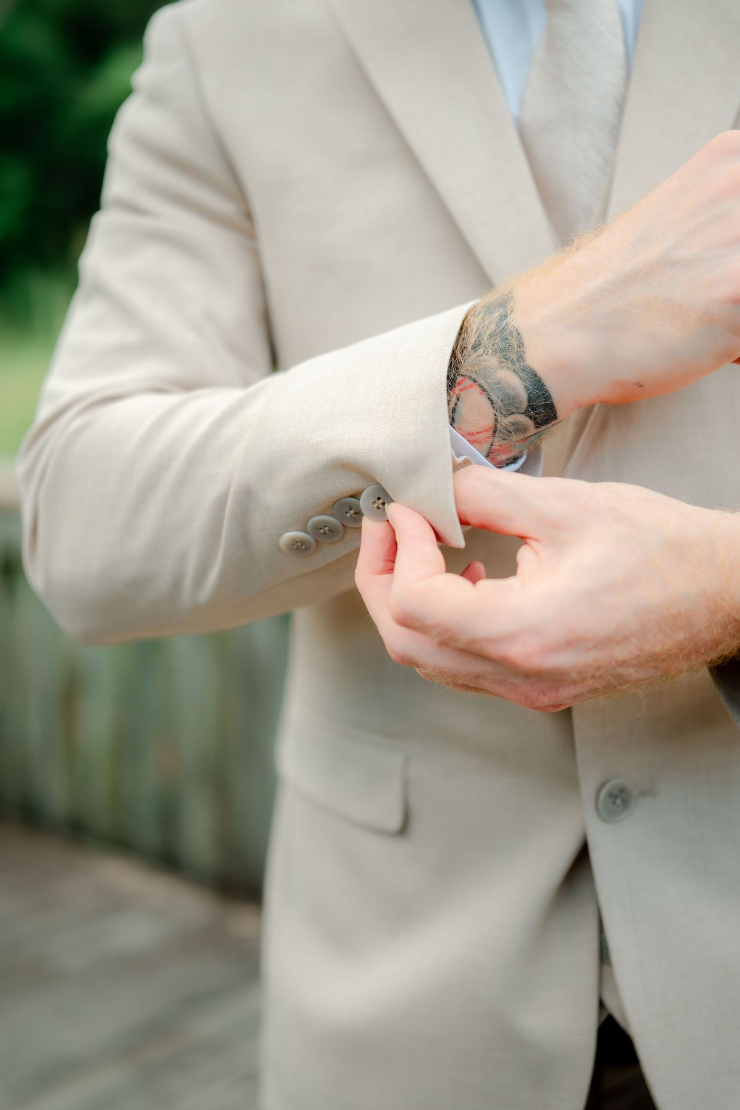 Close-up of someone adjusting the cuff of a light beige suit jacket, showing a glimpse of their tattoo artwork beneath.
