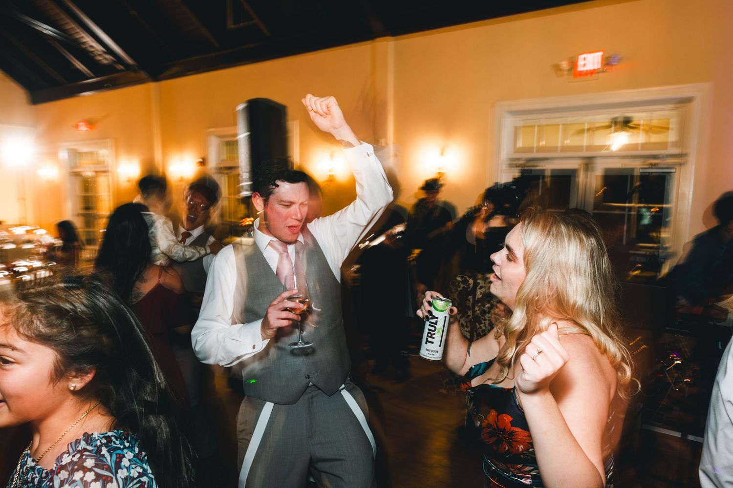 People dancing and celebrating at an indoor wedding reception with warm lighting and drinks in hand.