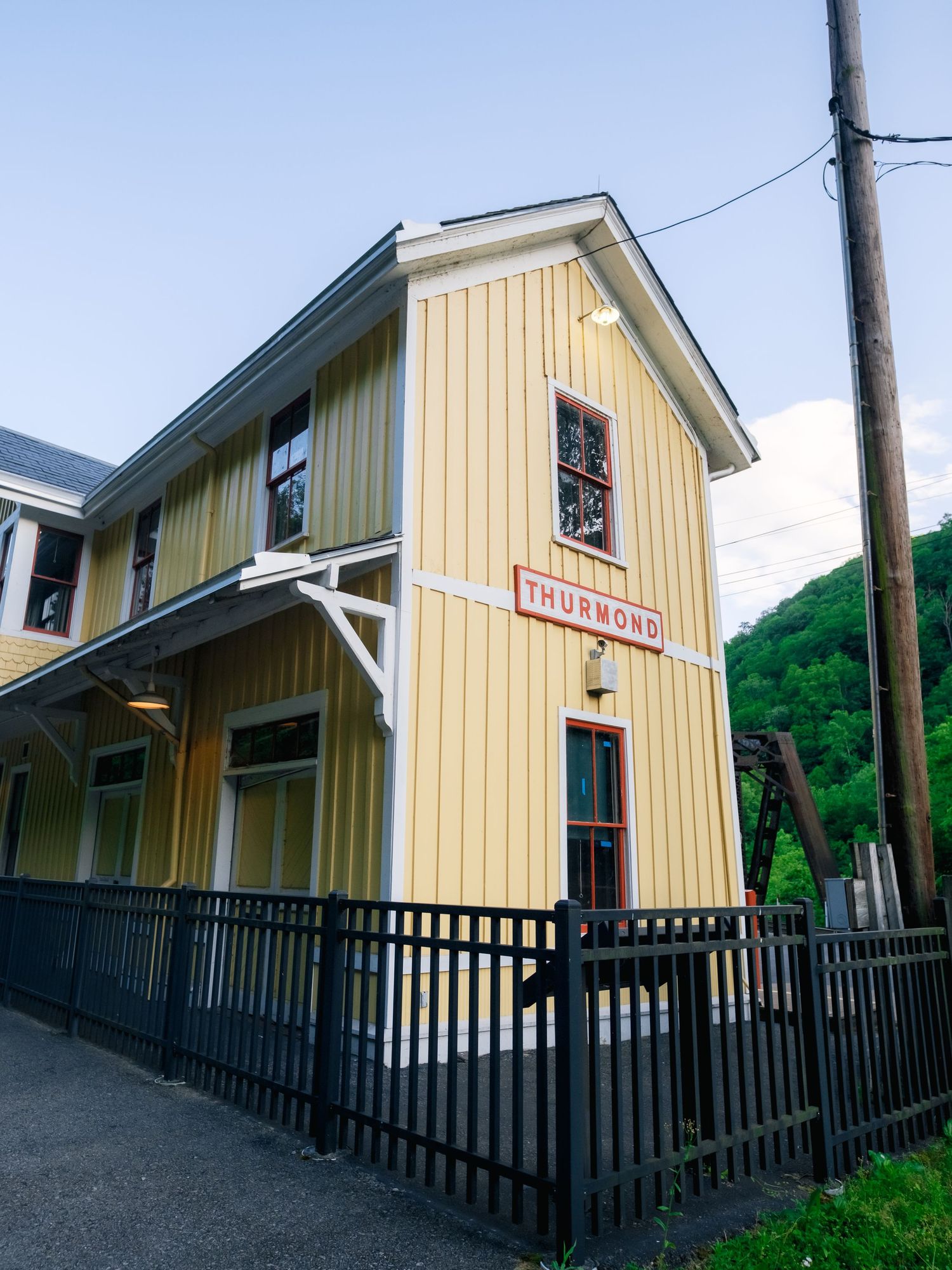 Vintage yellow train depot station with vertical siding and black railings against a blue sky and green hillside.