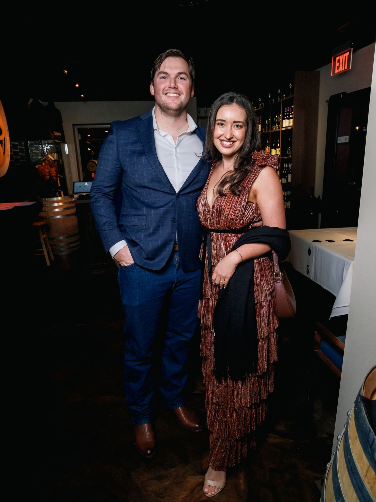 A couple in evening attire poses together at a dimly lit indoor venue with dark ambient lighting.