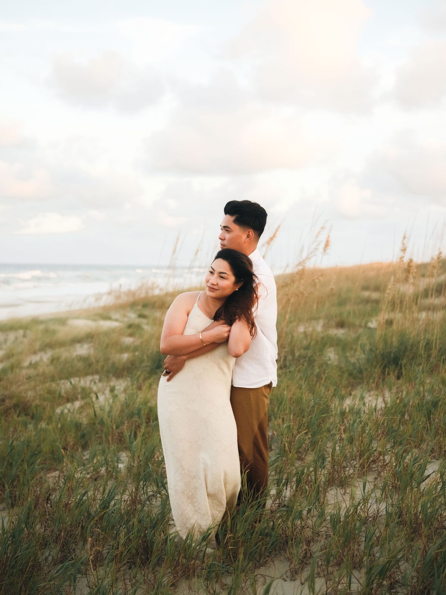 Couple embracing in flowing grass field during golden hour with cloudy sky backdrop.