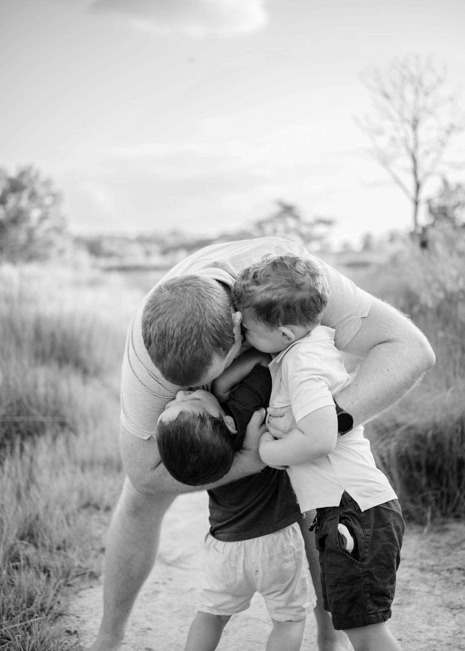 A touching black and white photo of a family embrace on a path surrounded by tall grass and trees in nature.