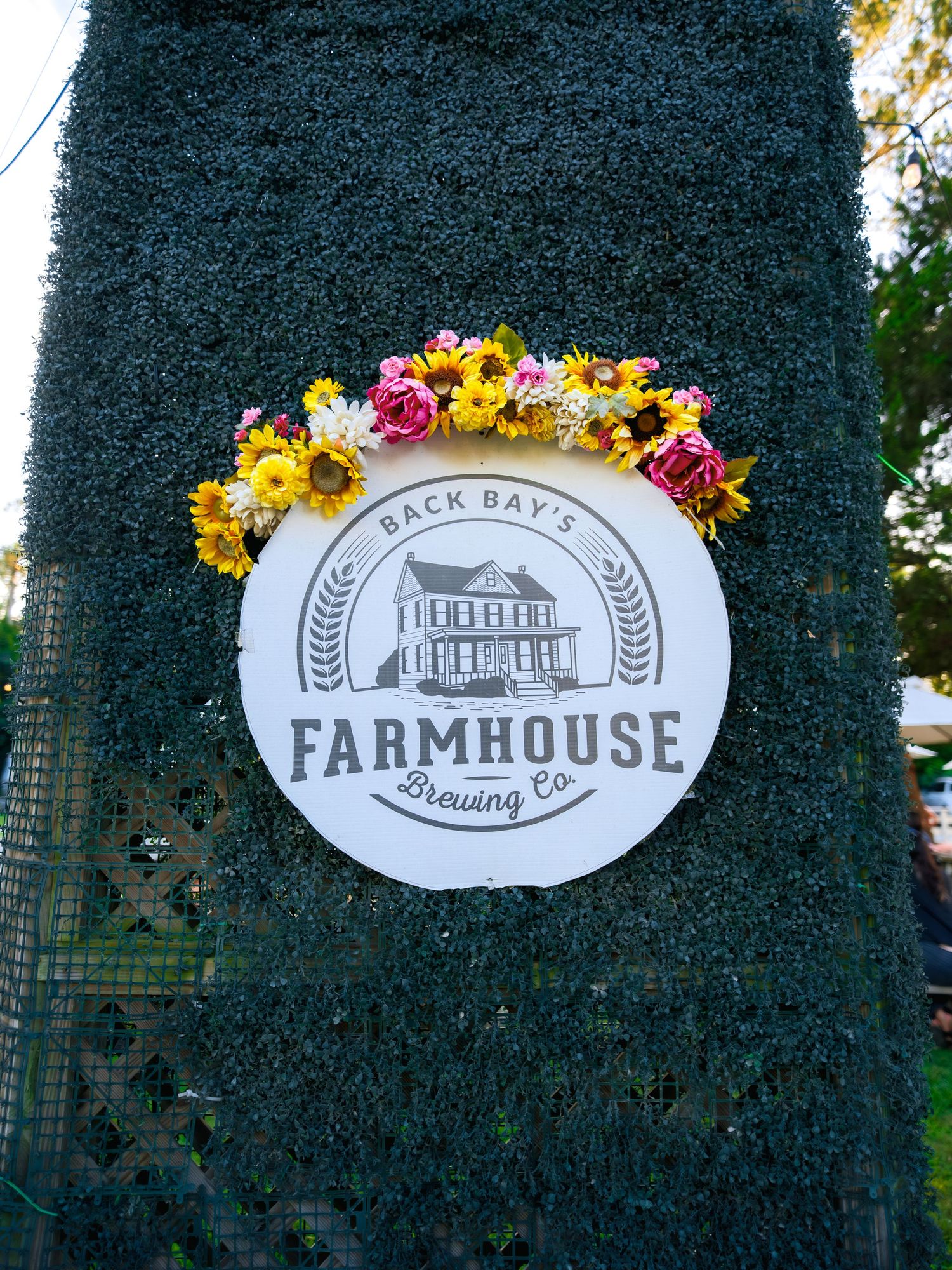 White circular Farmhouse sign adorned with colorful fresh flowers including yellow and purple blooms against dark background.