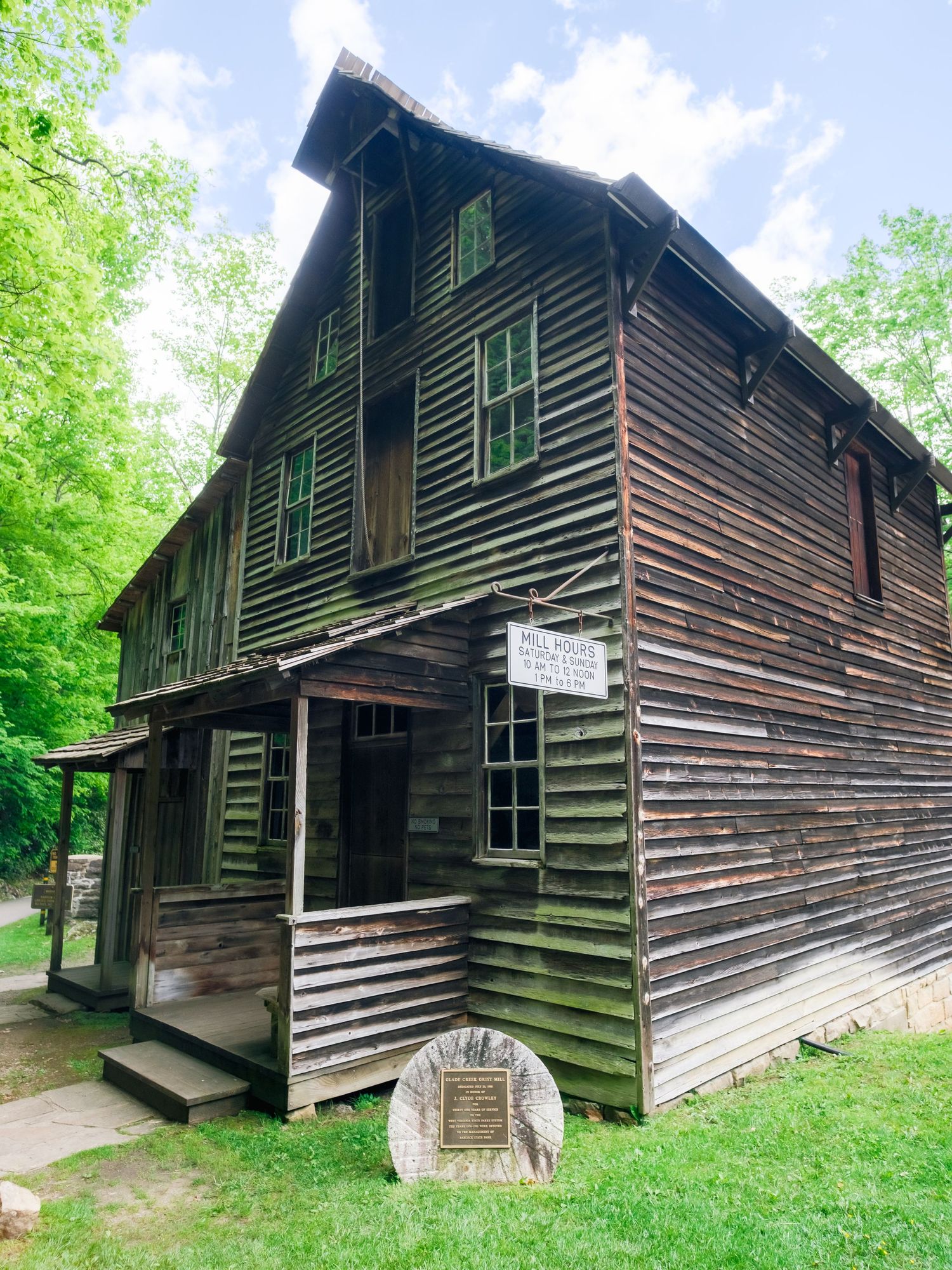 Historic wooden mill or general store building with weathered siding and stone marker stands among green trees on grassy ground.